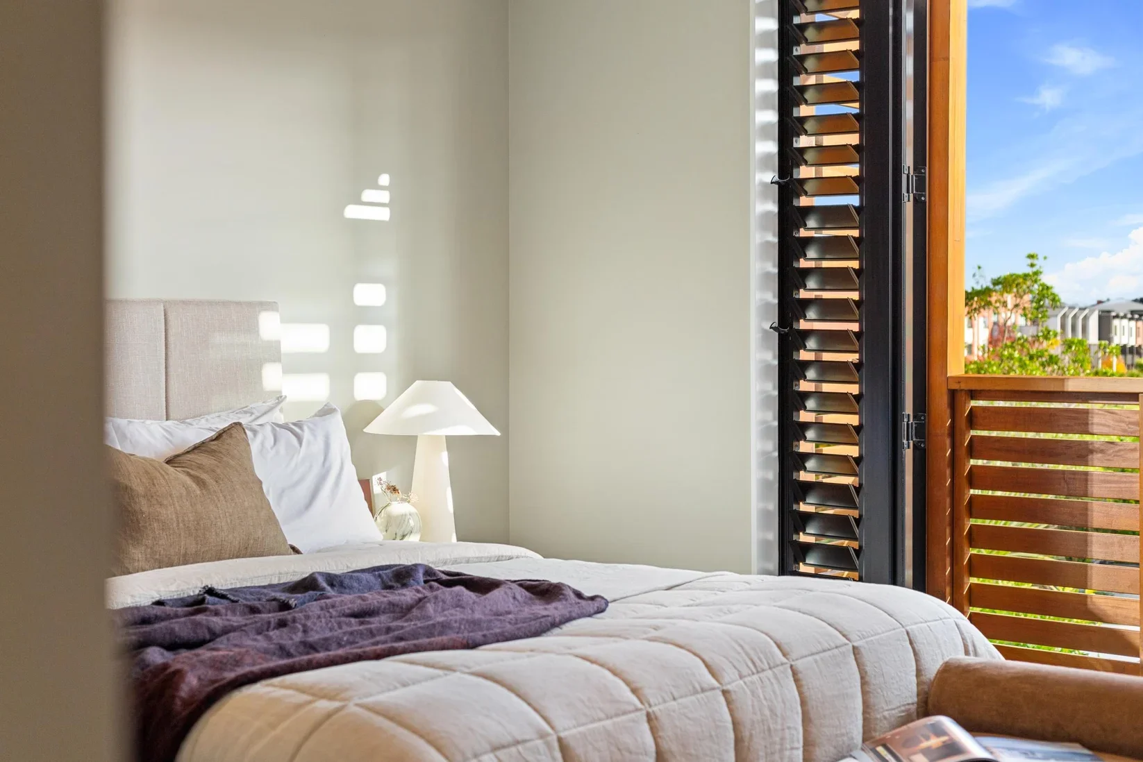 A neatly made bed with beige and white pillows, a beige headboard, and a purple blanket. A white bedside lamp and a glass vase with dried flowers are on a nightstand. The room has a large window with wooden blinds, sunlight casting shadows on the light green wall, and a view of trees and neighboring buildings outside.