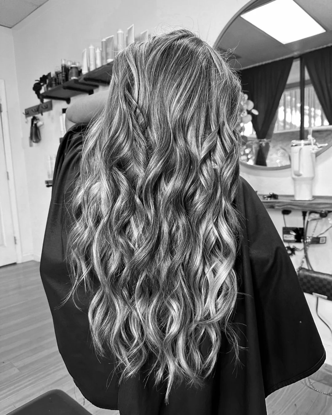 A woman with long, wavy hair sitting in a salon chair. Her hair is styled with loose curls. The salon has shelves with hair products, a mirror, and a large window with curtains.