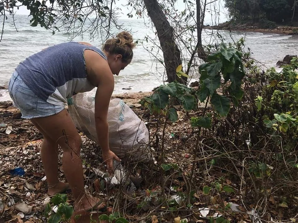 Person collecting trash in a beach area surrounded by trees and plants.
