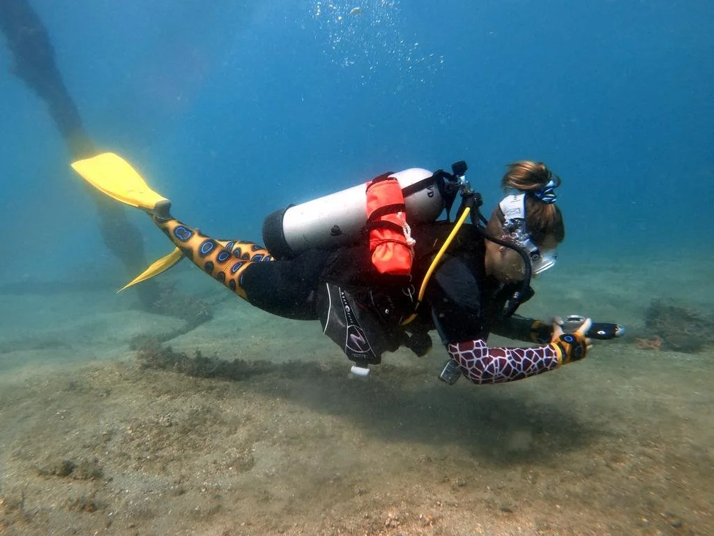 Person in scuba diving gear, with colorful fish-patterned tail and arms, using an underwater camera, exploring the ocean floor.