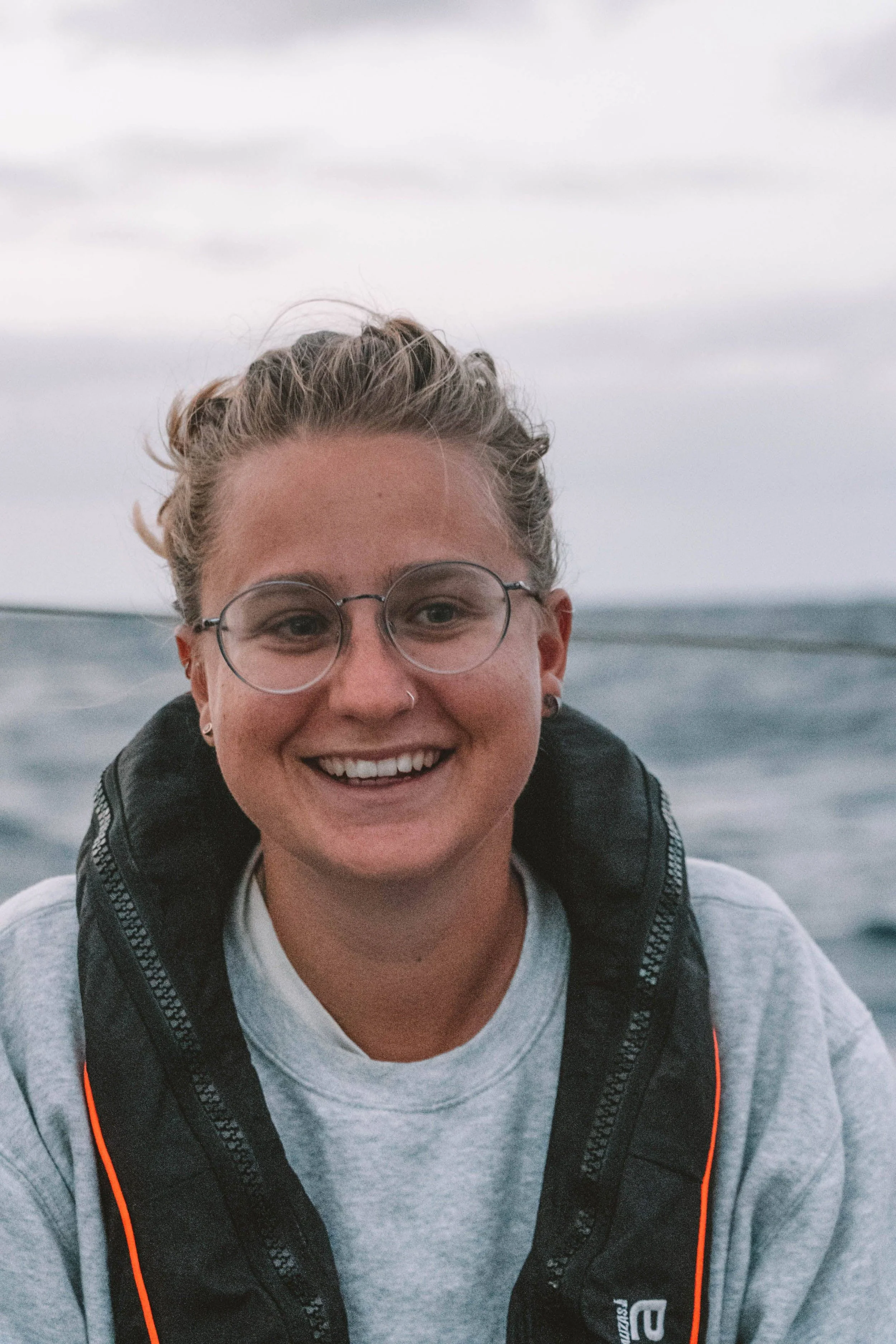 A young woman with curly blonde hair in a bun, wearing glasses, a nose ring, a gray sweatshirt, and a black life jacket, smiling on a boat with water and a cloudy sky in the background.