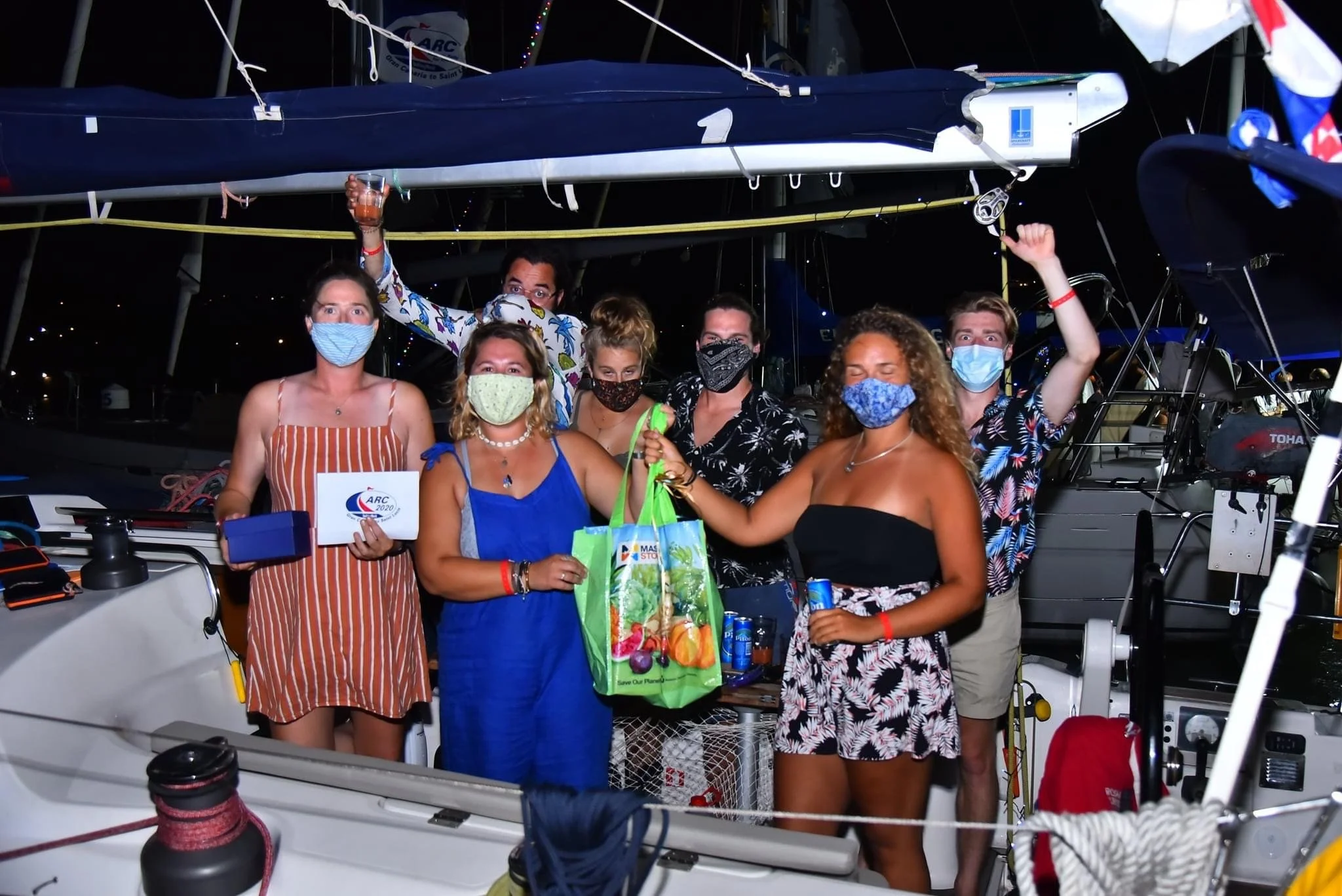 Group of people on a boat celebrating, wearing masks, with some holding drinks and gifts, at night with other boats in the background.