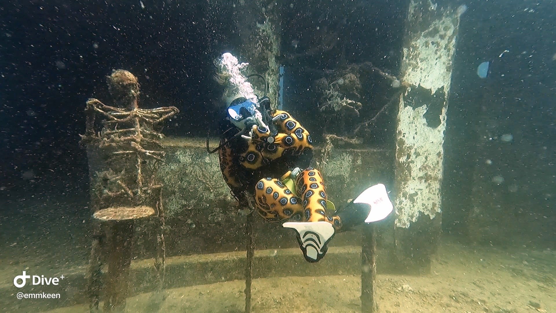 A scuba diver wearing an orange suit with blue and black eyes and white fins inspects an underwater statue of a skeleton leaning on a chainsaw, with another skeleton behind it, on the ocean floor.