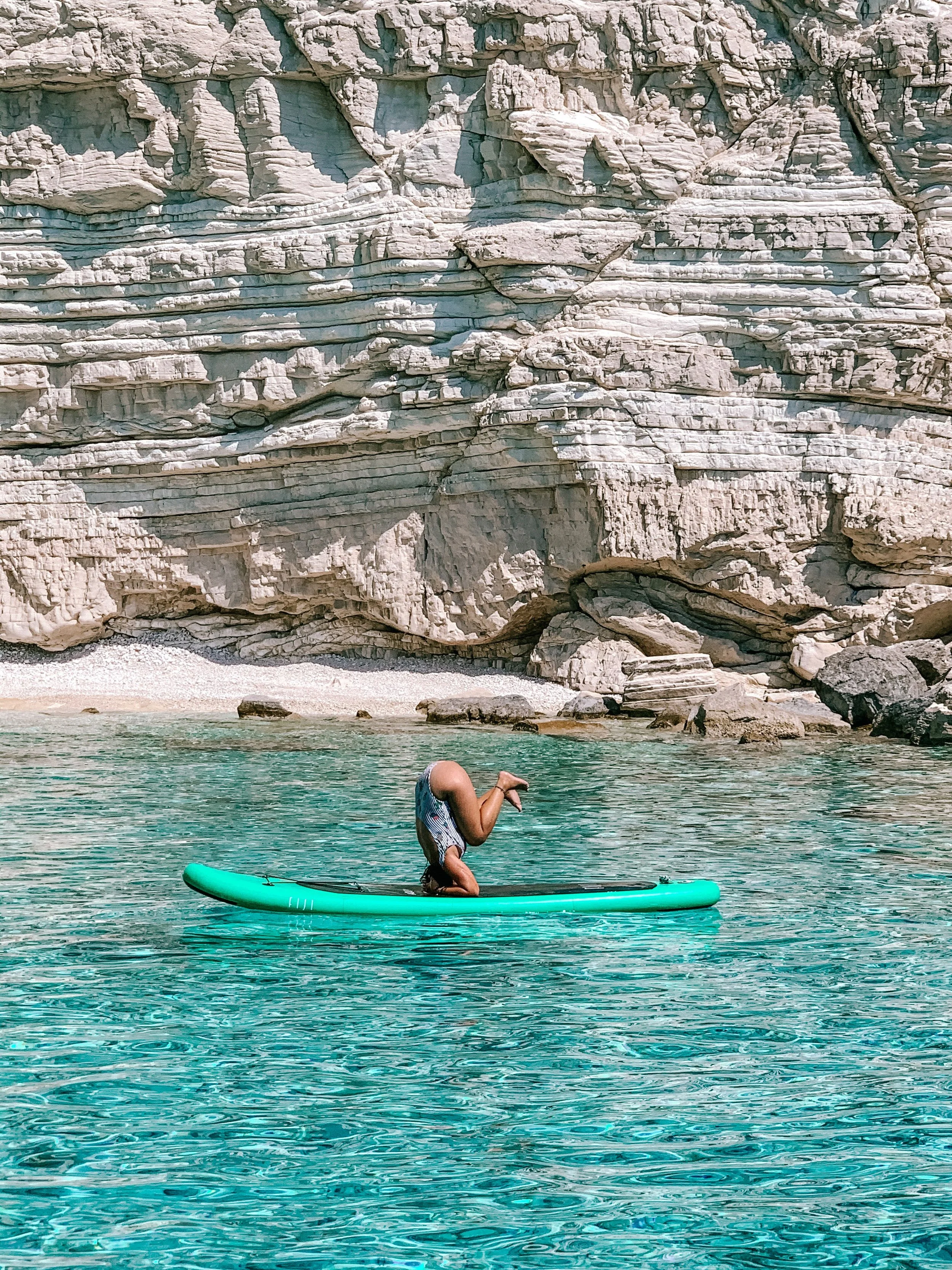 Person doing a headstand on a paddleboard in a clear, turquoise body of water with a rocky, cliffside background.
