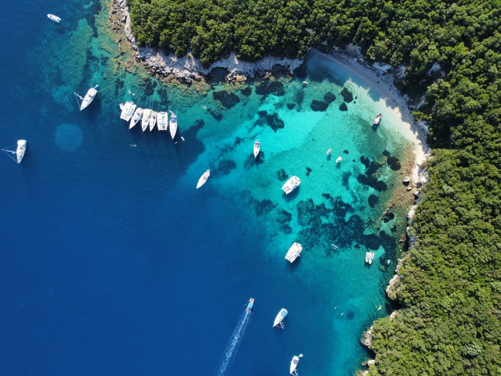 An aerial view of a coastal area with turquoise water, several boats anchored near a forested shoreline, and a small beach with people relaxing.