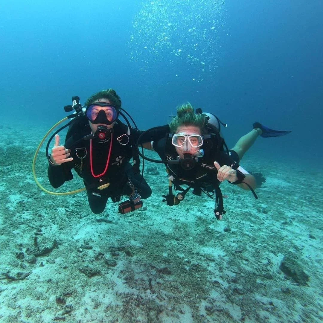 Two scuba divers underwater on the ocean floor, wearing wetsuits, masks, and oxygen tanks, giving thumbs up and making the shaka sign.