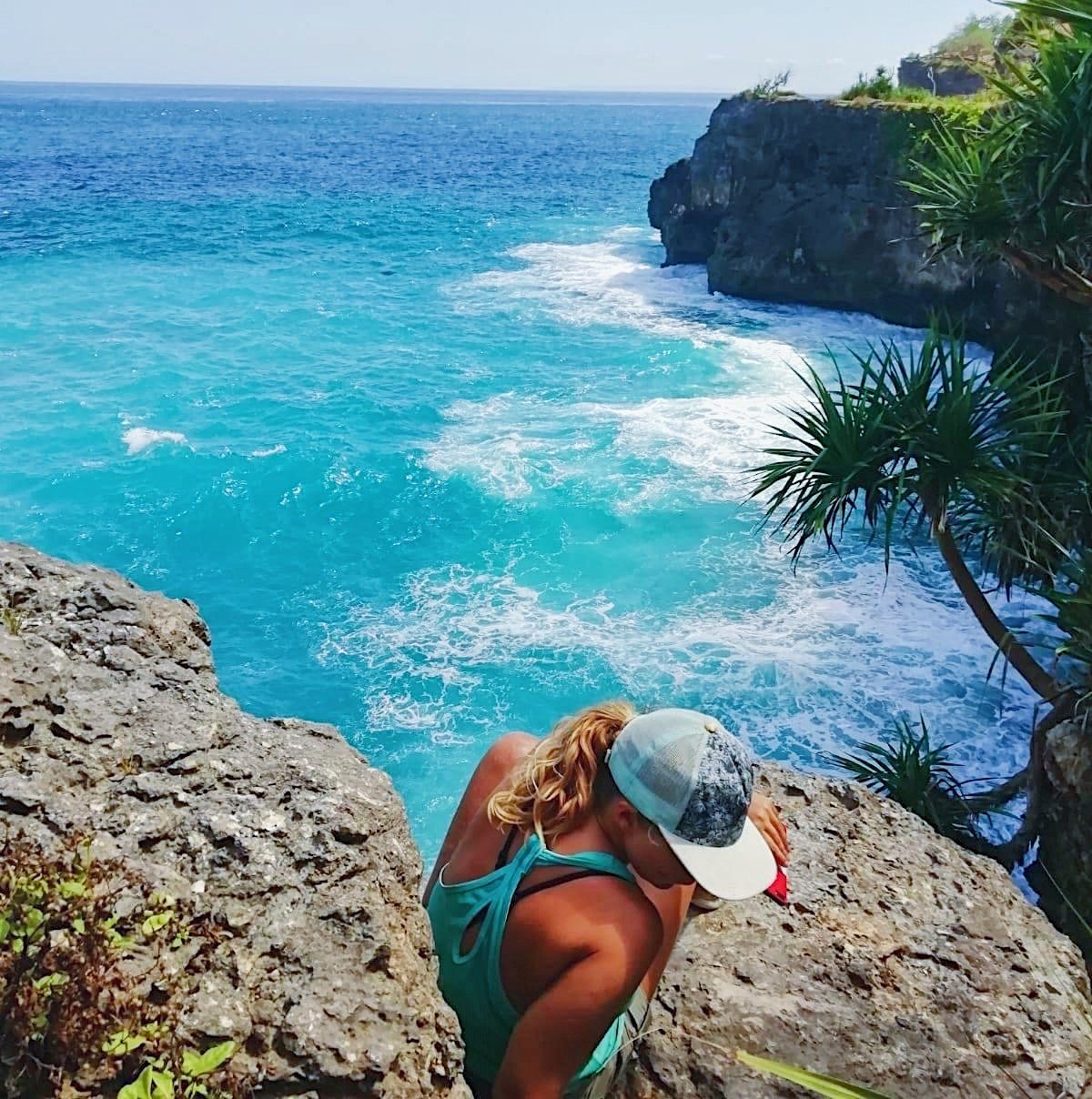 A person with blonde hair wearing a cap and turquoise tank top crouches on a rocky cliff overlooking the ocean, with turquoise water, a distant horizon, and some greenery.
