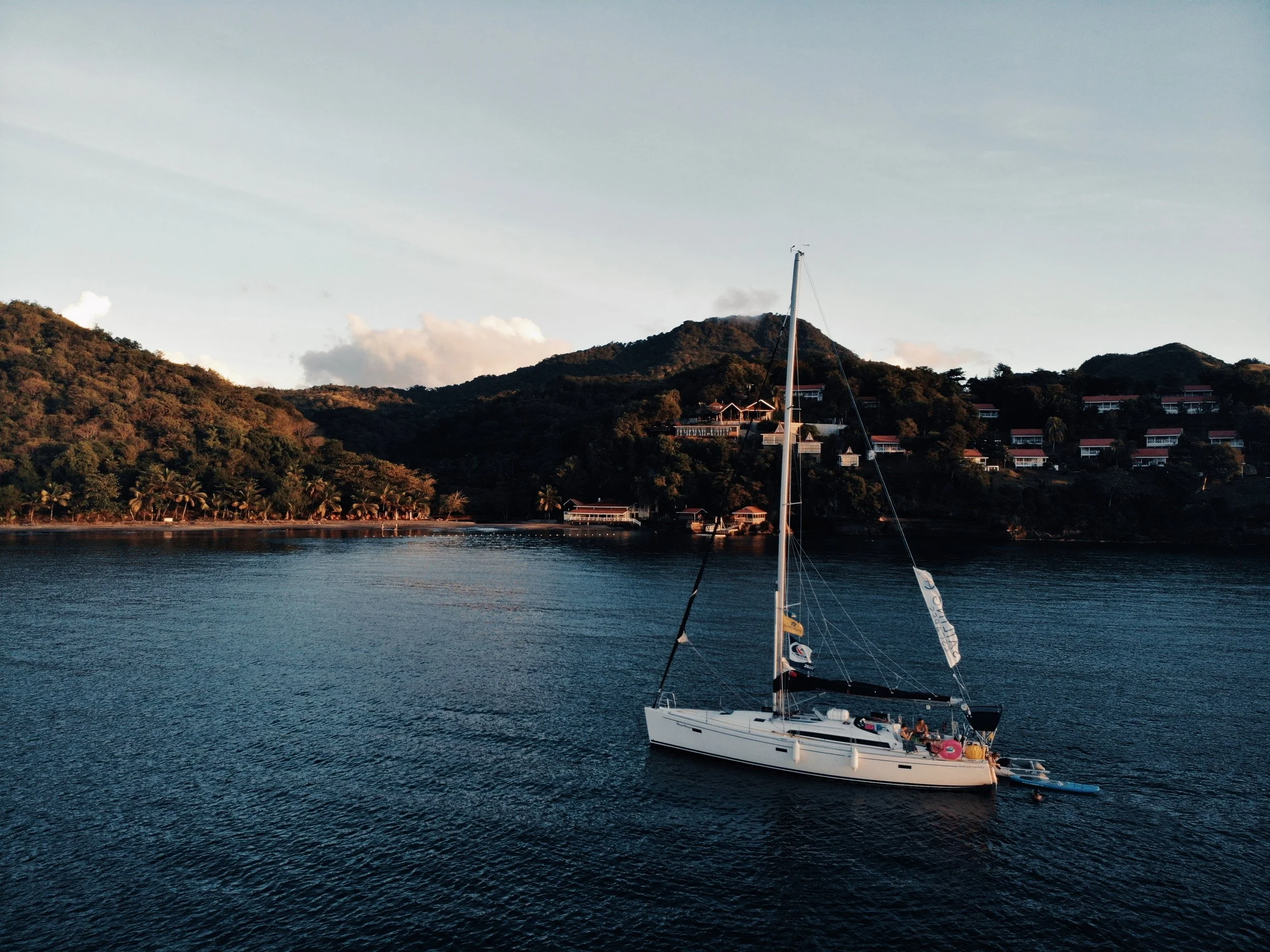 A sailboat with a sail and small motor attached, floating on calm water near a lush, hilly shoreline with buildings and trees, under a partly cloudy sky at sunset.