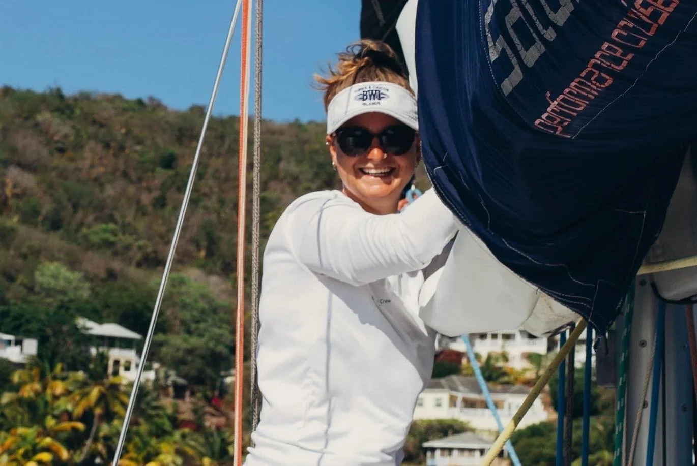 Woman smiling, wearing sunglasses and a white visor, adjusting sail on a sailboat, with boats and hillside houses in the background.