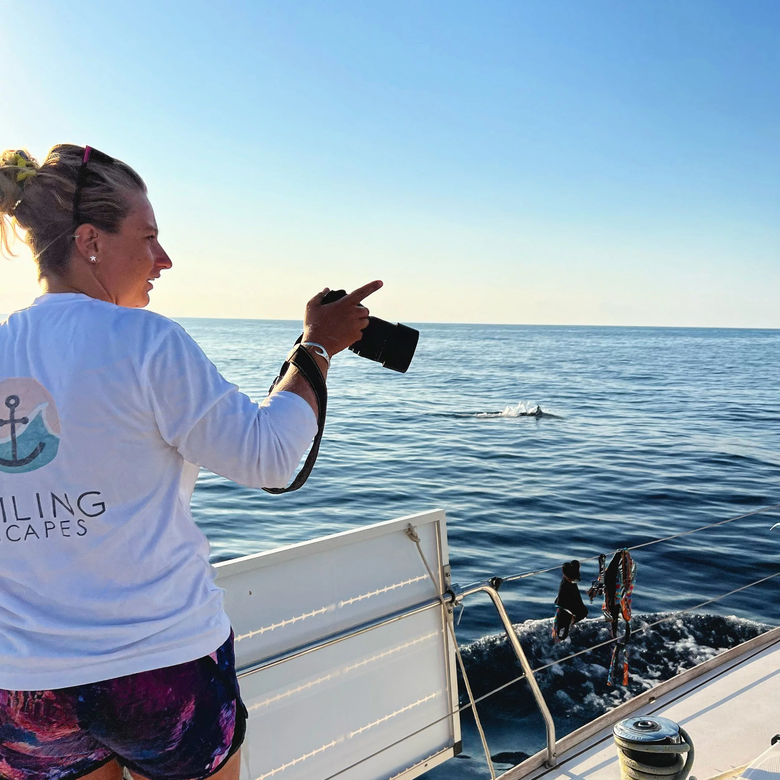 A woman on a boat taking a photo or video of a whale swimming in the ocean during sunset.
