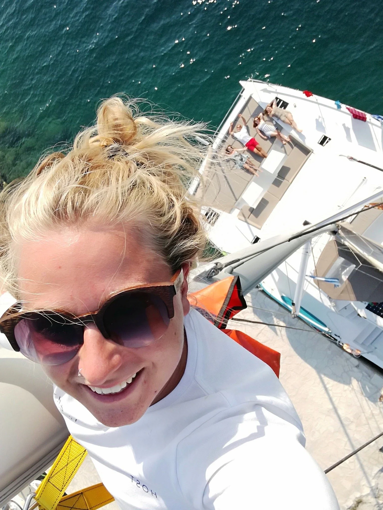 Person with blonde hair and sunglasses taking a selfie on a boat with people lounging on the deck in the background.