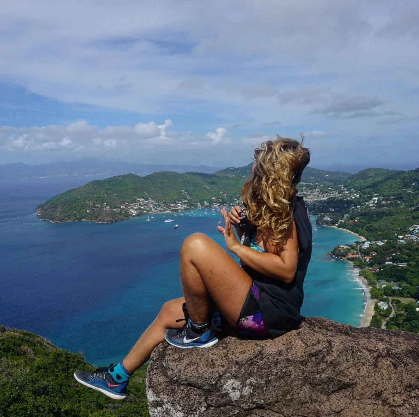 Person with curly hair sitting on a rock at a high vantage point overlooking a coastal landscape with mountains, trees, water, and a beach.
