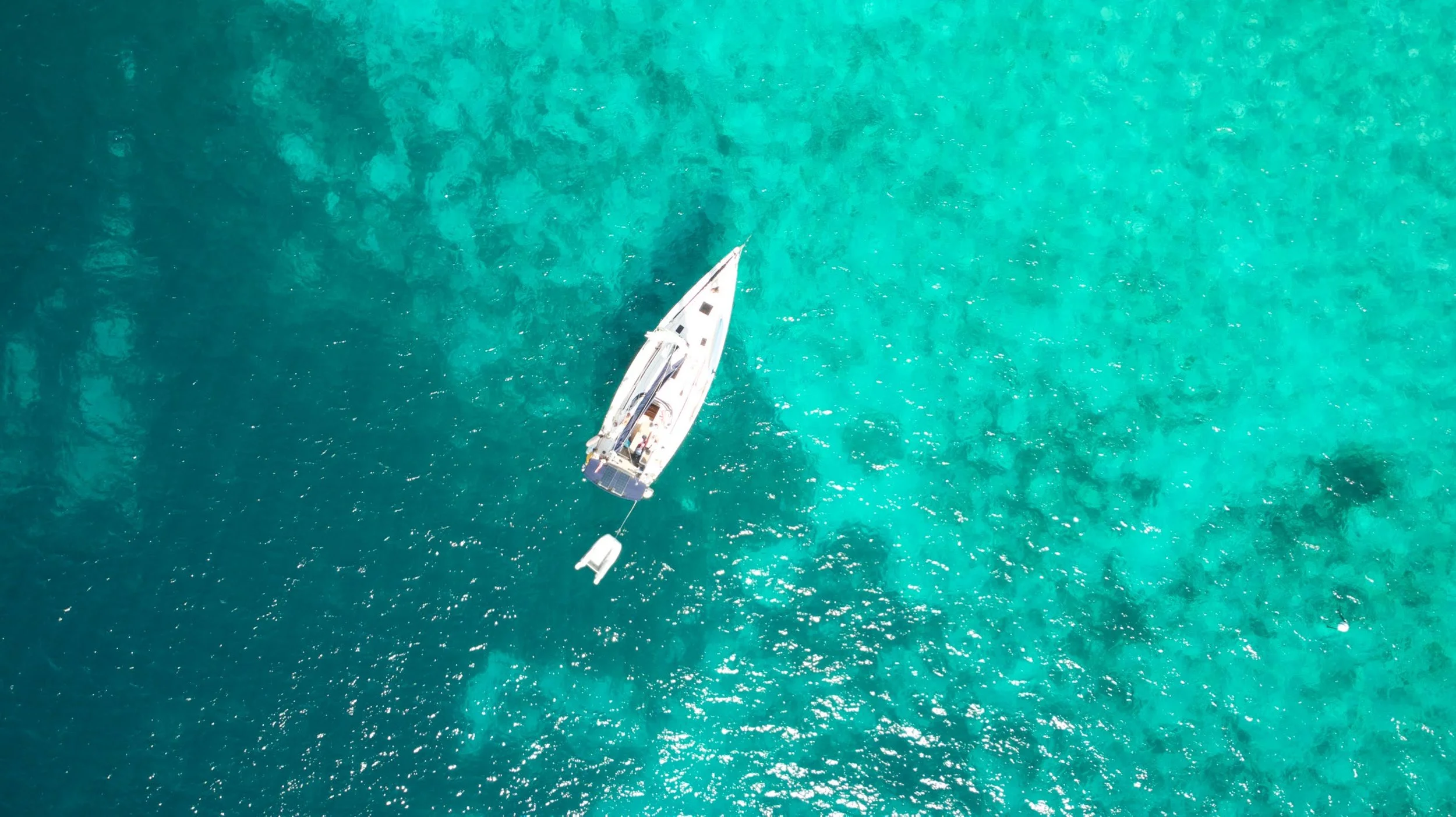 An aerial view of a sailboat anchored in turquoise water with coral reef visible below the surface.