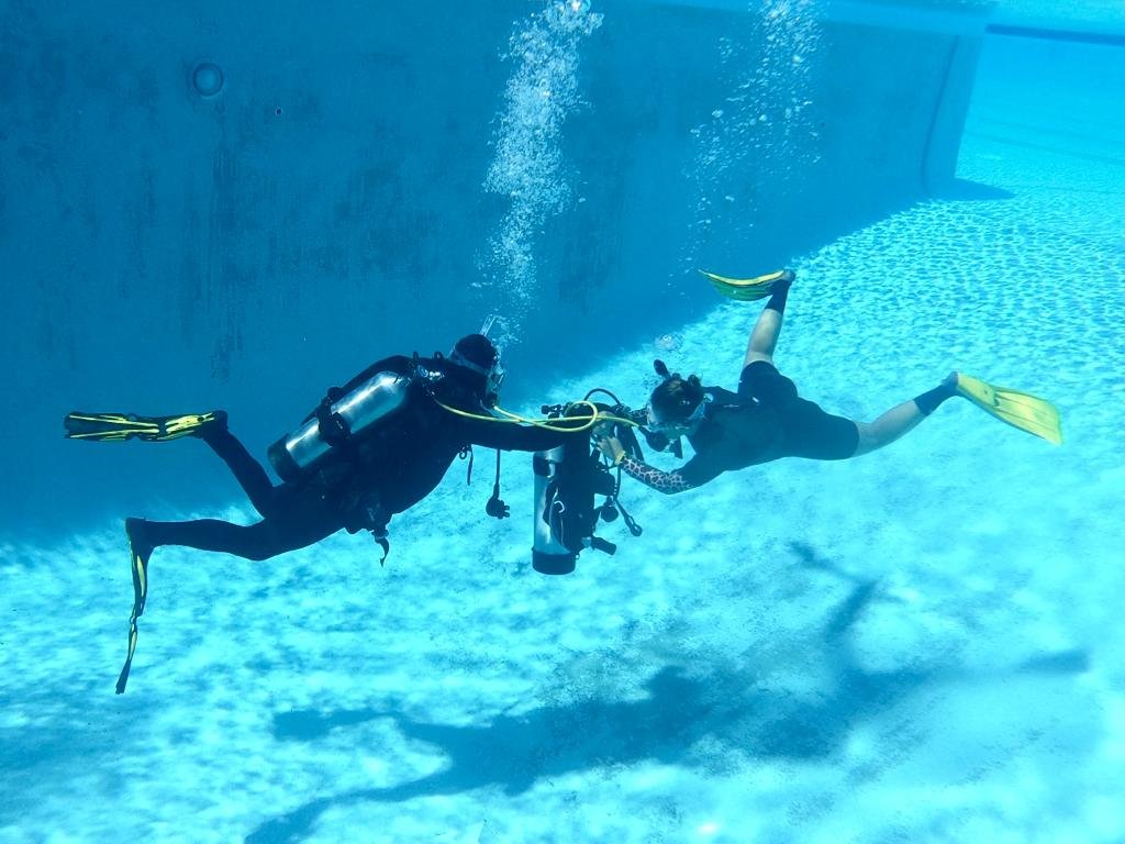 Two scuba divers swimming underwater near a pool wall, holding hands and surrounded by clear blue water.