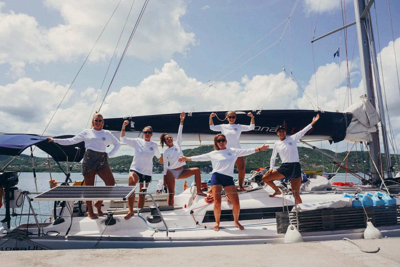 Six women in white shirts posing on a sailboat docked at a marina with green hills in the background under a partly cloudy sky.