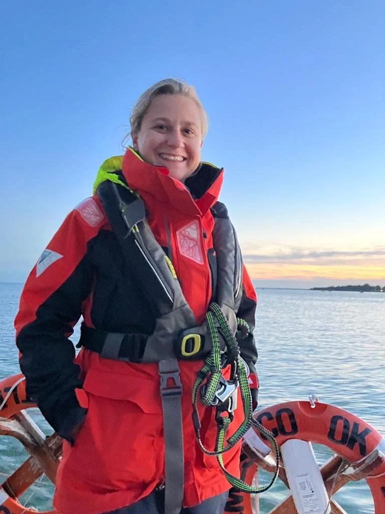 A woman in a red and black life jacket and red waterproof gear standing on a boat, smiling, with water and a sunset sky in the background.