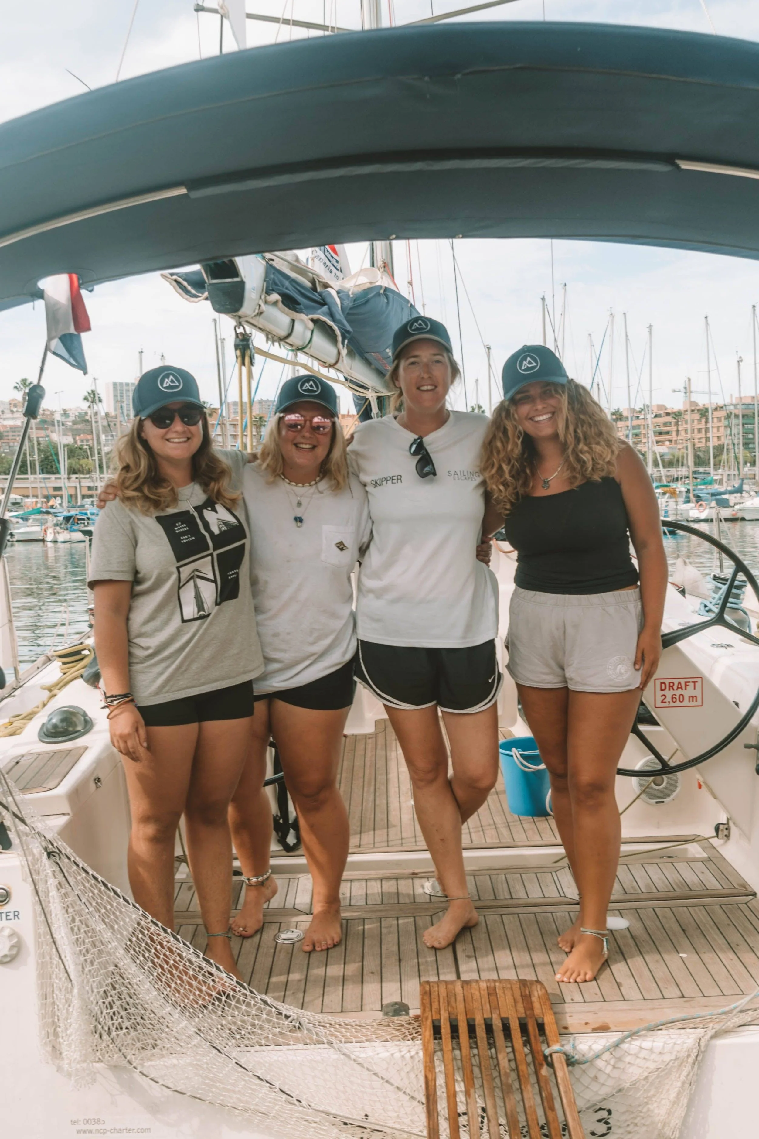 Four women standing on a sailboat dock, smiling. They are wearing casual clothing and sailing caps. The background shows marina with sailboats and buildings.