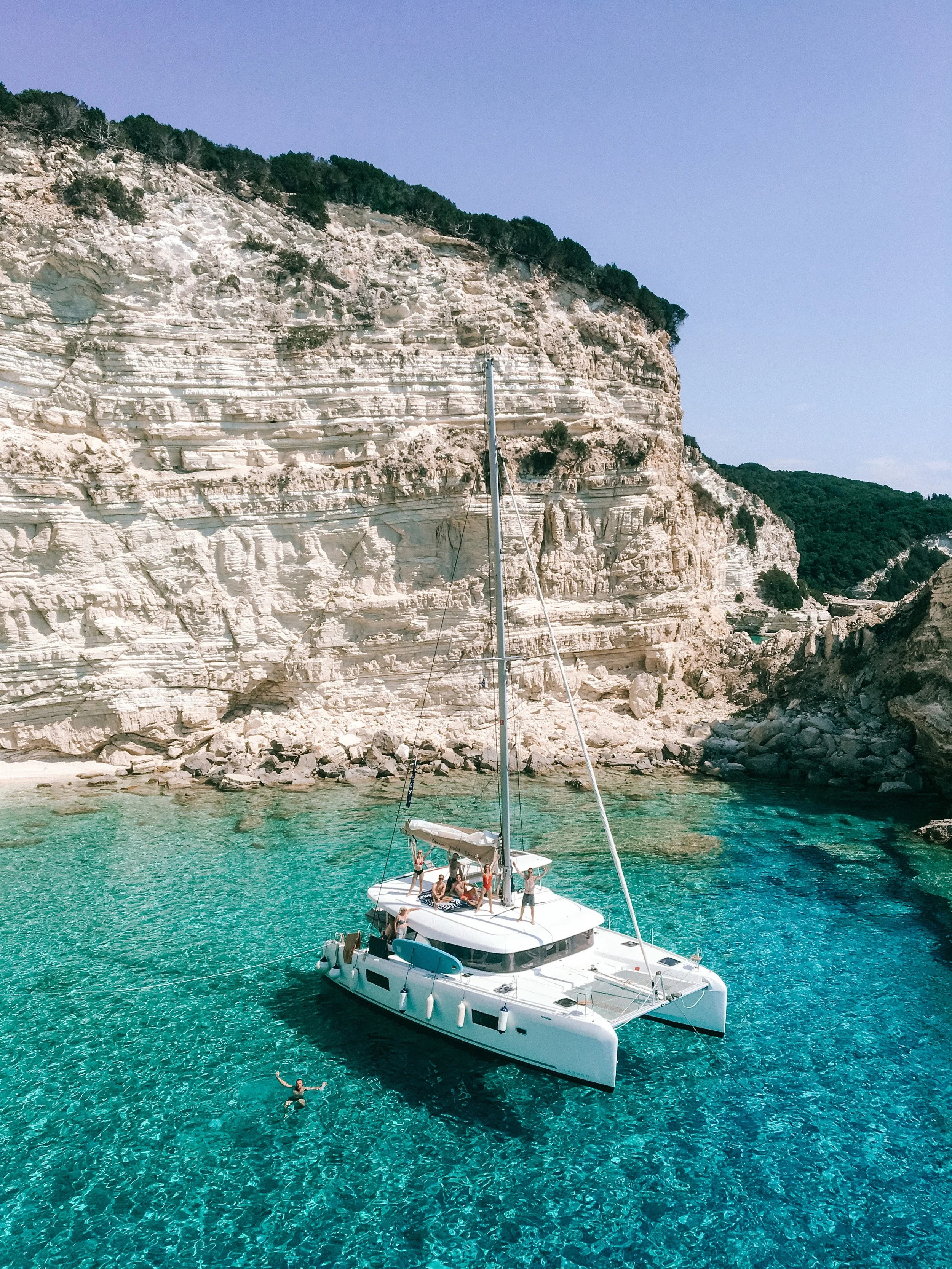 A white catamaran boat with people onboard and one person swimming peacefully in clear turquoise water near rocky cliffs on a sunny day.