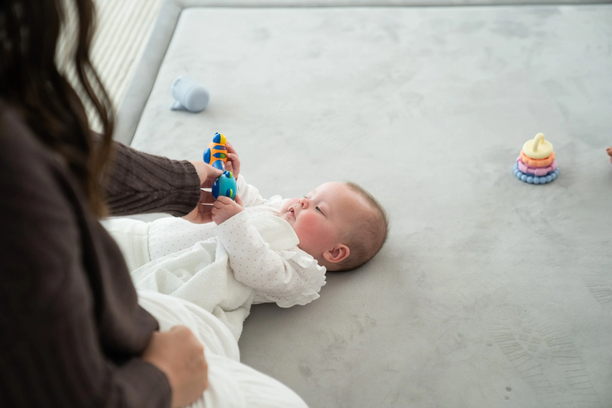 A baby lying on a light gray carpet, reaching for a toy held by an adult during a starting solids feeding workshop through Let's Start Solids Adelaide