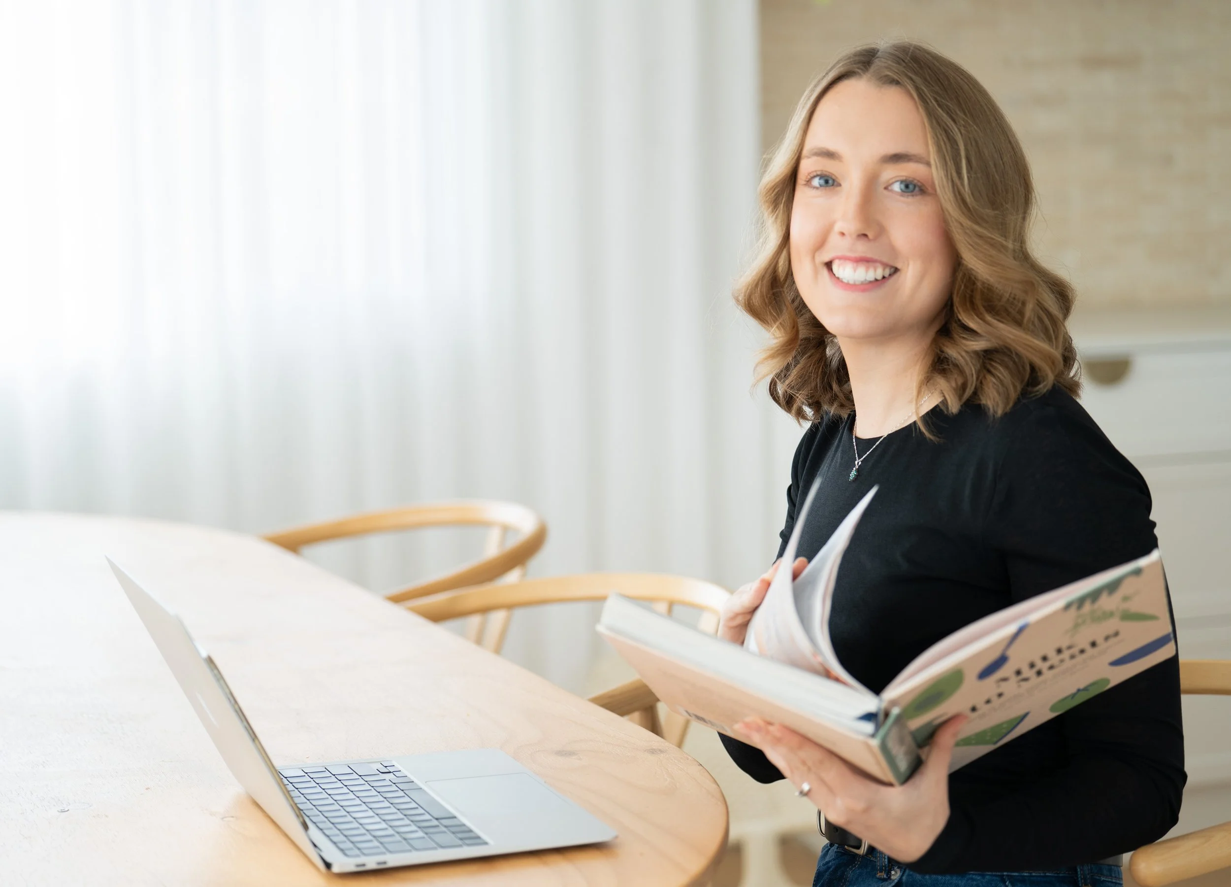 Healthcare expert and feeding therapist in Adelaide with wavy blonde hair smiling, sitting at a desk holding an open book, with a laptop open in front of her, running starting solids consultations and workshops through Let's Start Solids