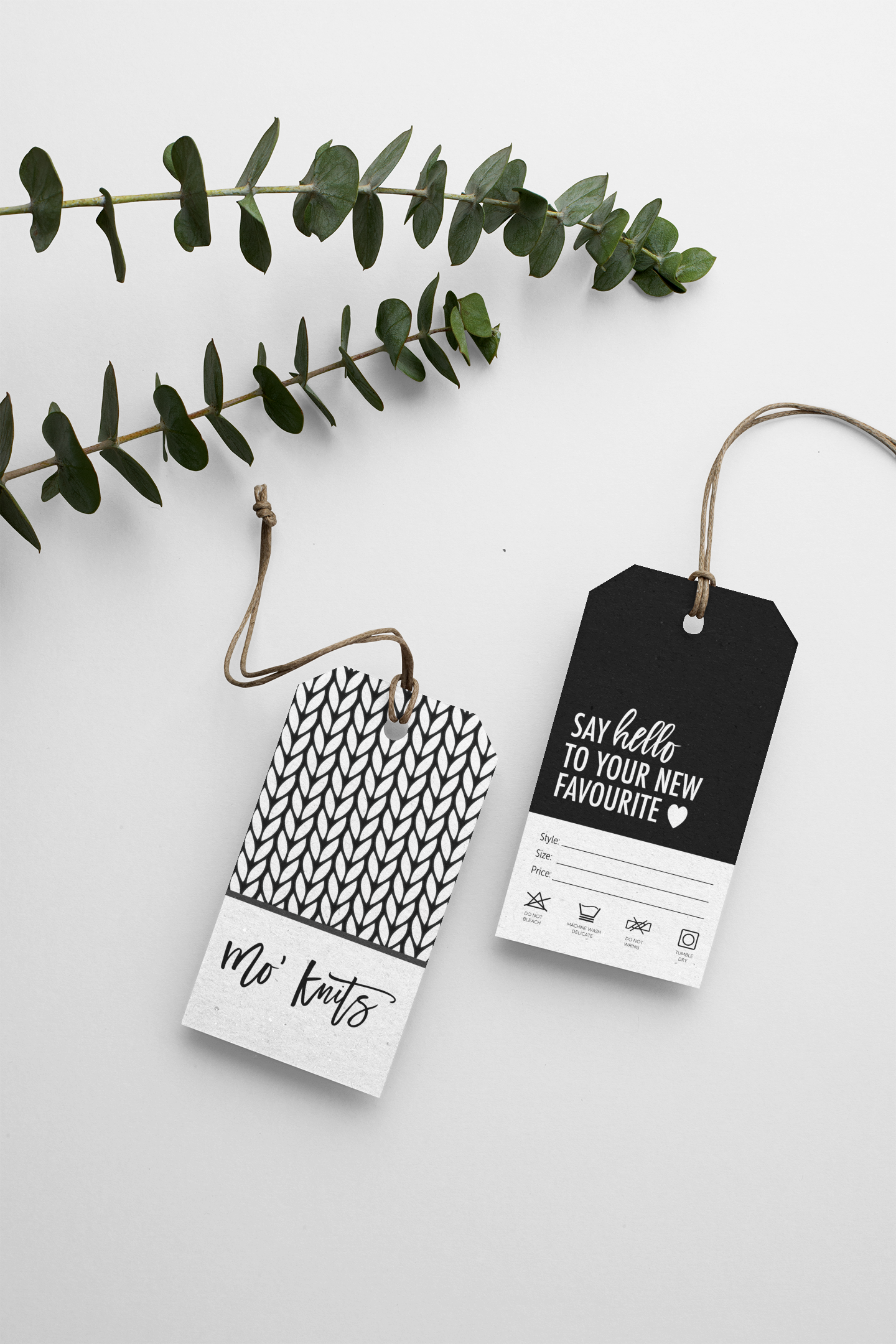 Two clothing tags, one with a black and white leaf pattern and the text 'Mo' Knits,' and the other black with white text saying 'Say hello to your new favorite,' placed on a white surface next to a green eucalyptus branch.