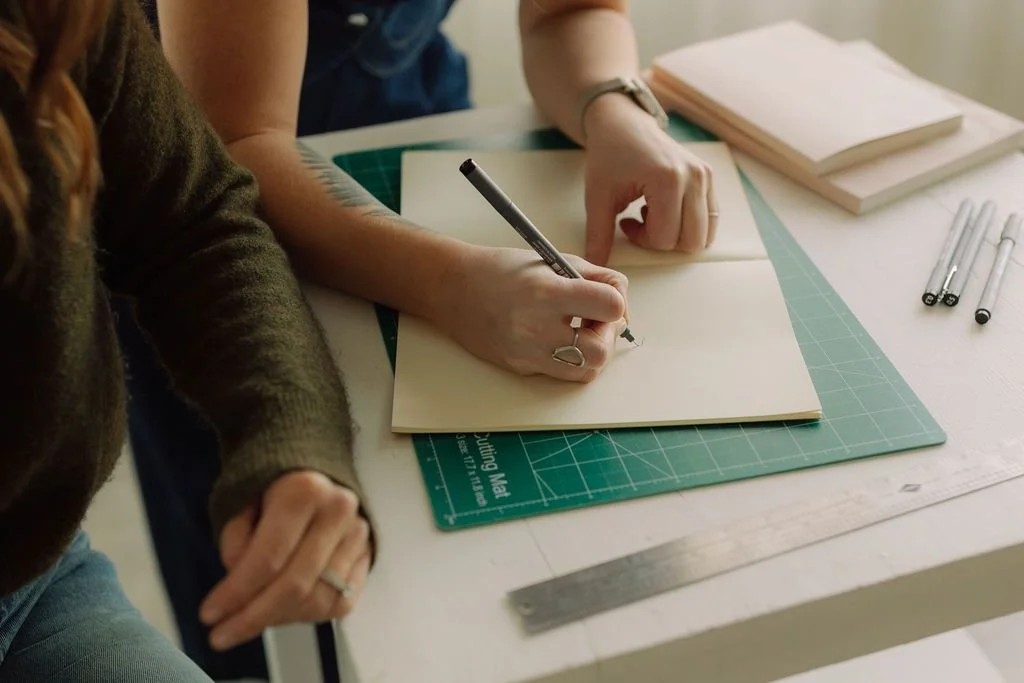 Person writing in an open notebook on a drafting table with drawing tools, markers, and stacked papers.