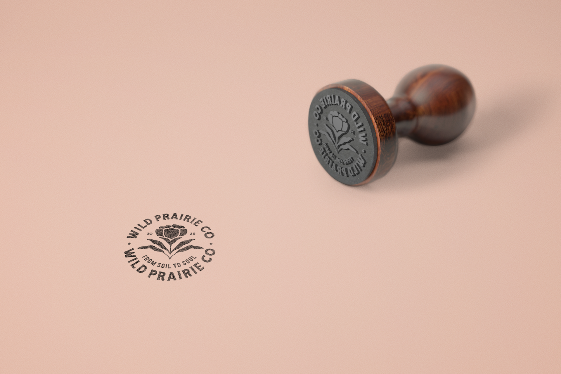 A rubber stamp with a wooden handle and a black logo on the stamp and on the pink surface below it. The logo features a flower with the text 'WILD PRAIRIE CO. FROM SOIL TO SOUL' around it.