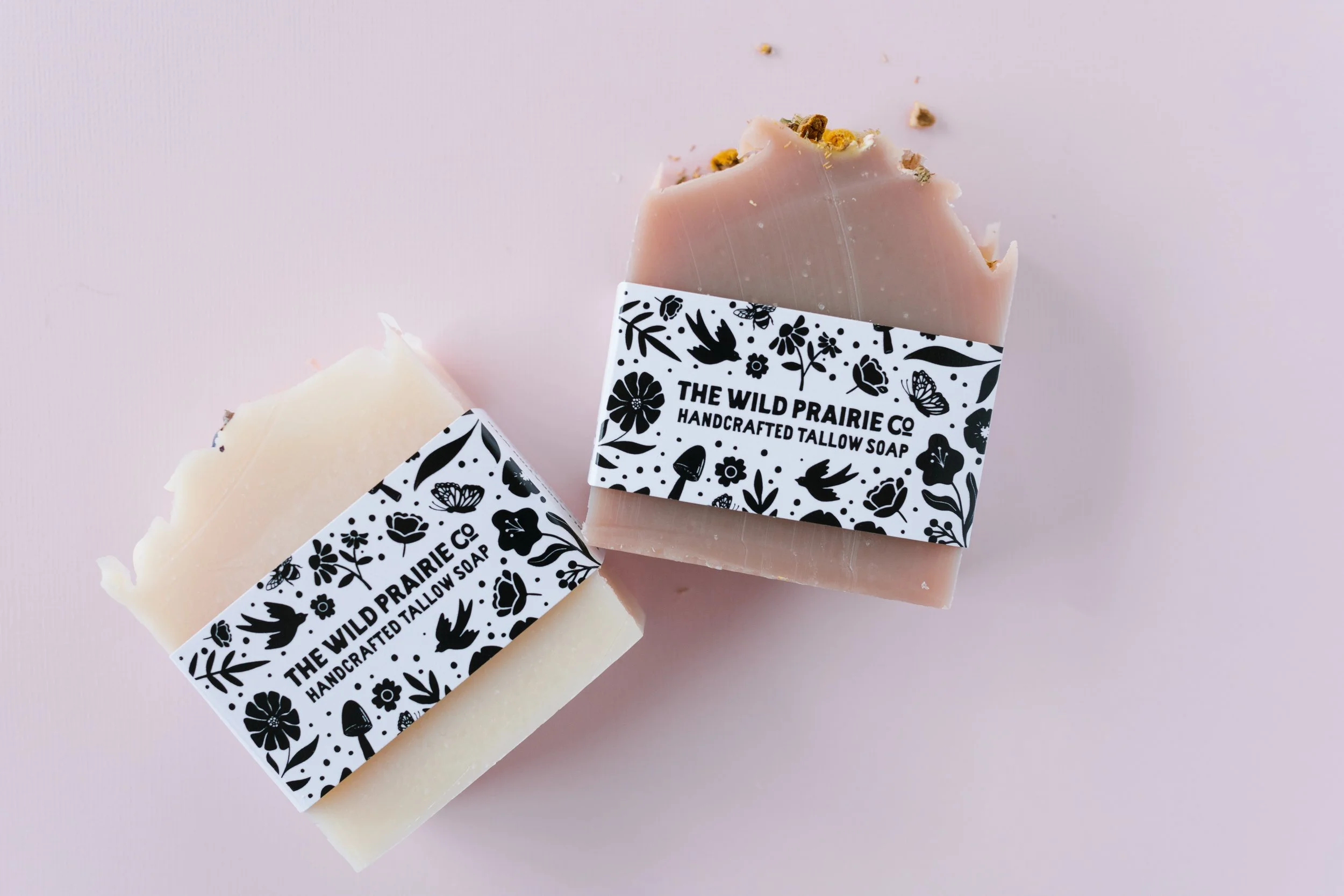 Two bars of handcrafted soap from The Wild Prairie Co, one plain cream-colored and one pink with flower and bird decorations, wrapped with black and white floral labels on a pink background.