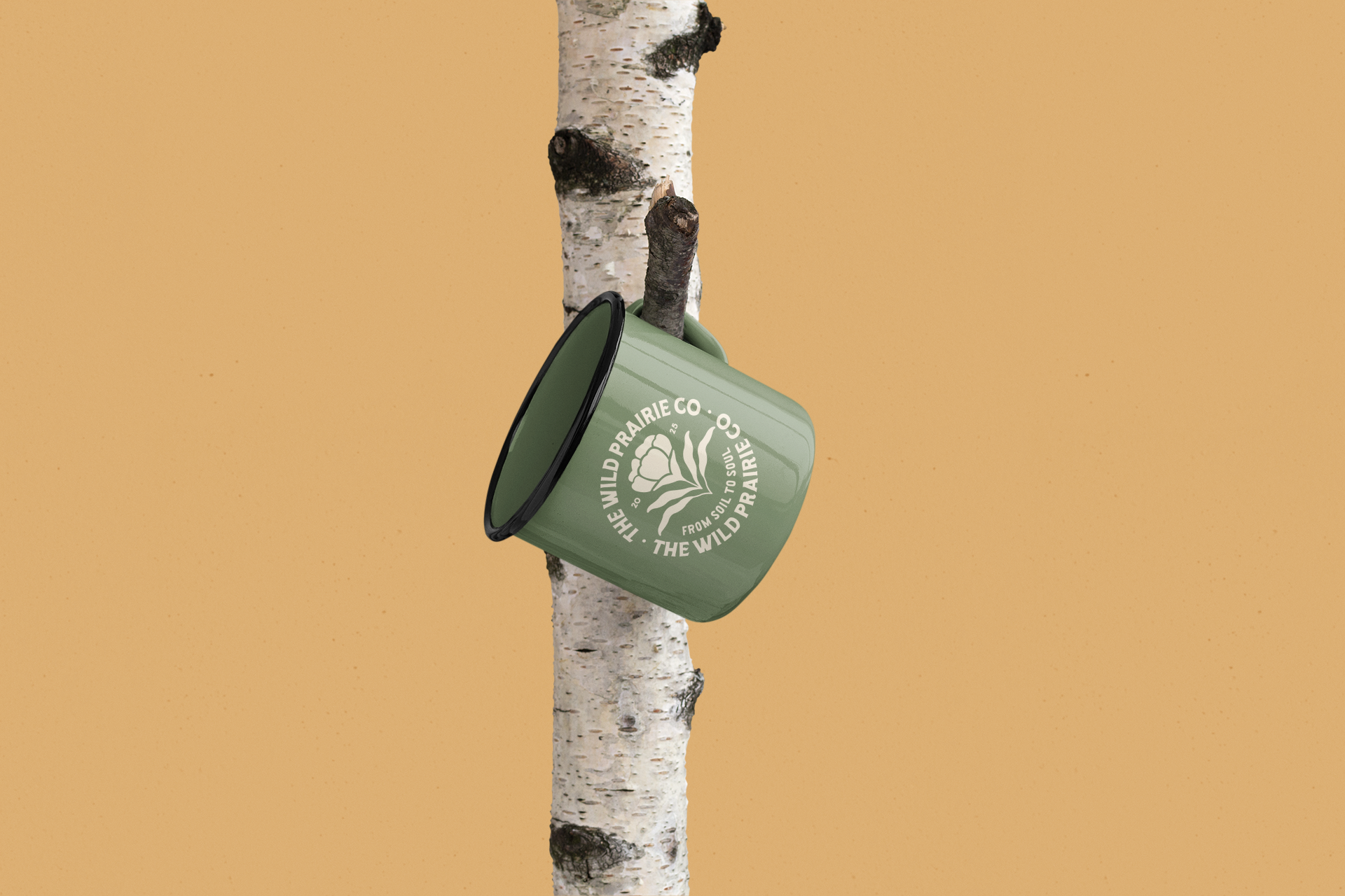 A green mug with white print labeled 'The Wild Prairie Co. from soil to s oil to soil' hanging on a small branch of a birch tree in front of a plain beige background.