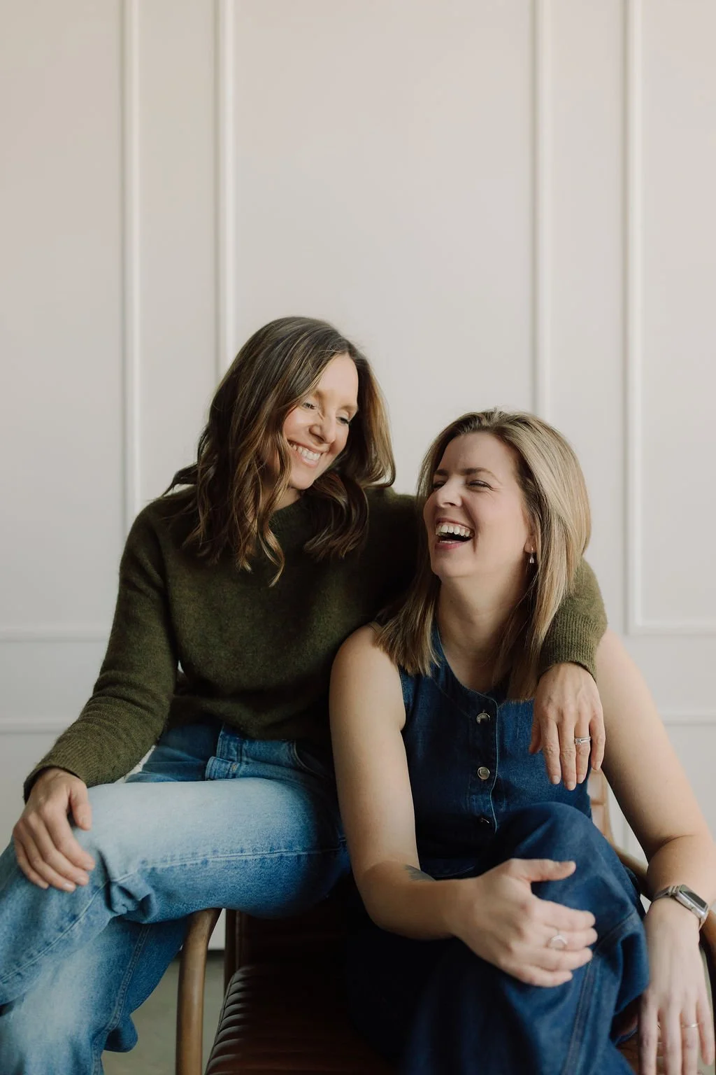 Jen & Janna  laughing and smiling, one with an arm around the other's shoulder, sitting together in a room with white paneled walls.