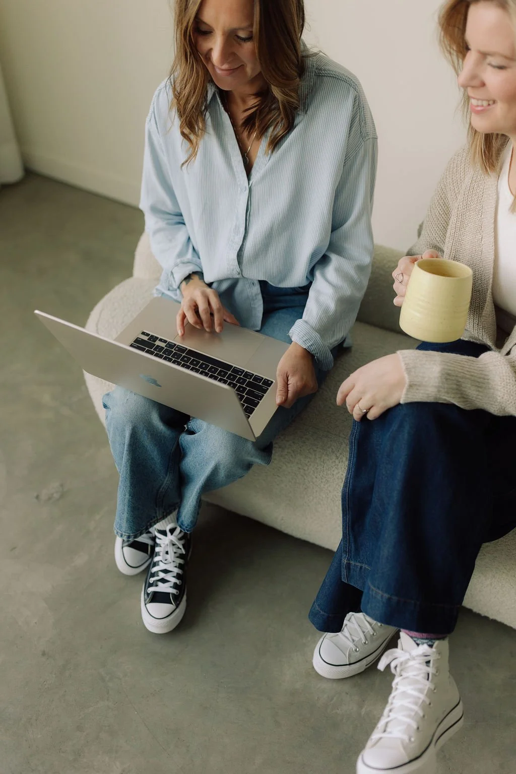 Two women sitting on a sofa, one using a laptop and the other holding a yellow mug, smiling and engaging in a conversation.