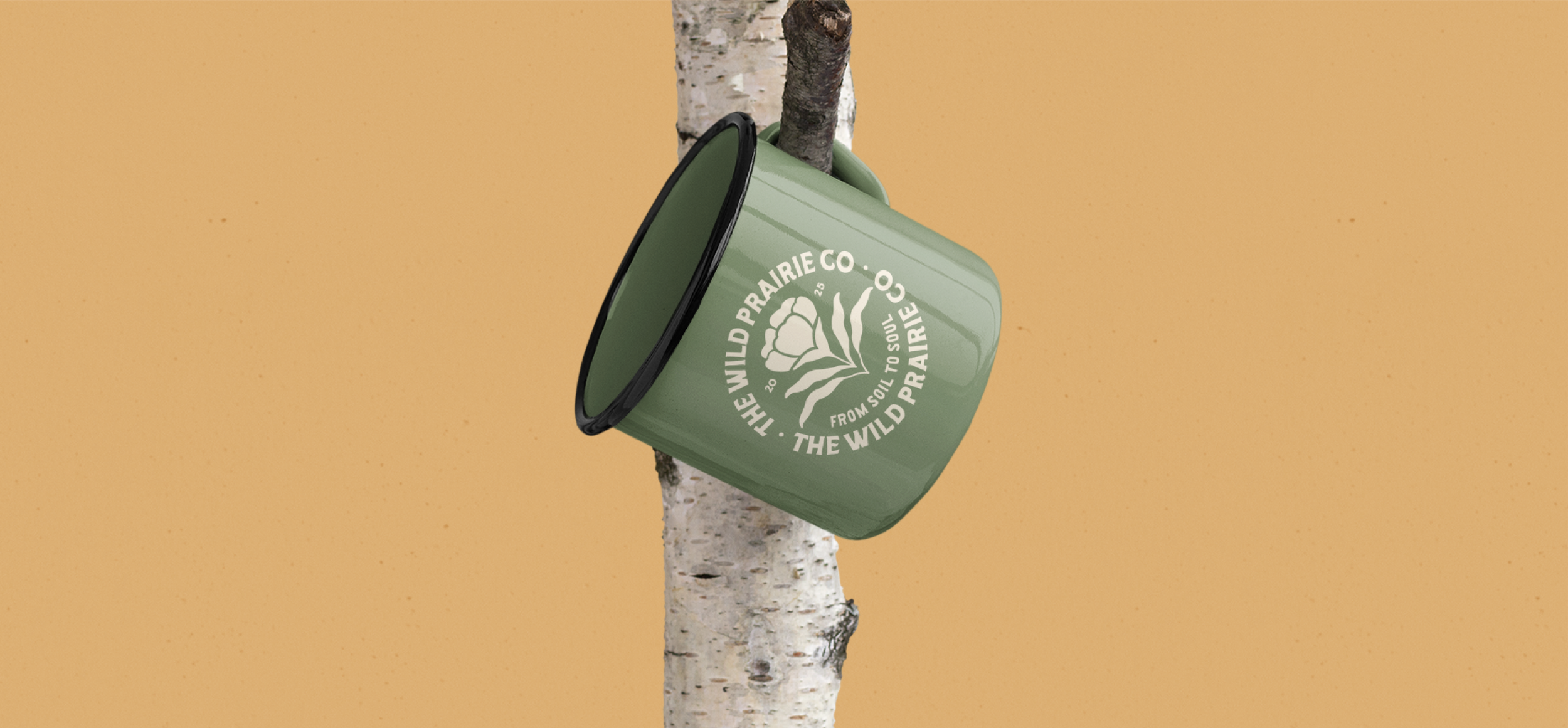 A green enamel camping mug with a white logo hanging on a tree trunk. The logo features a wildflower and reads "The Wild Prairie Co. - From Soil to Soul -".