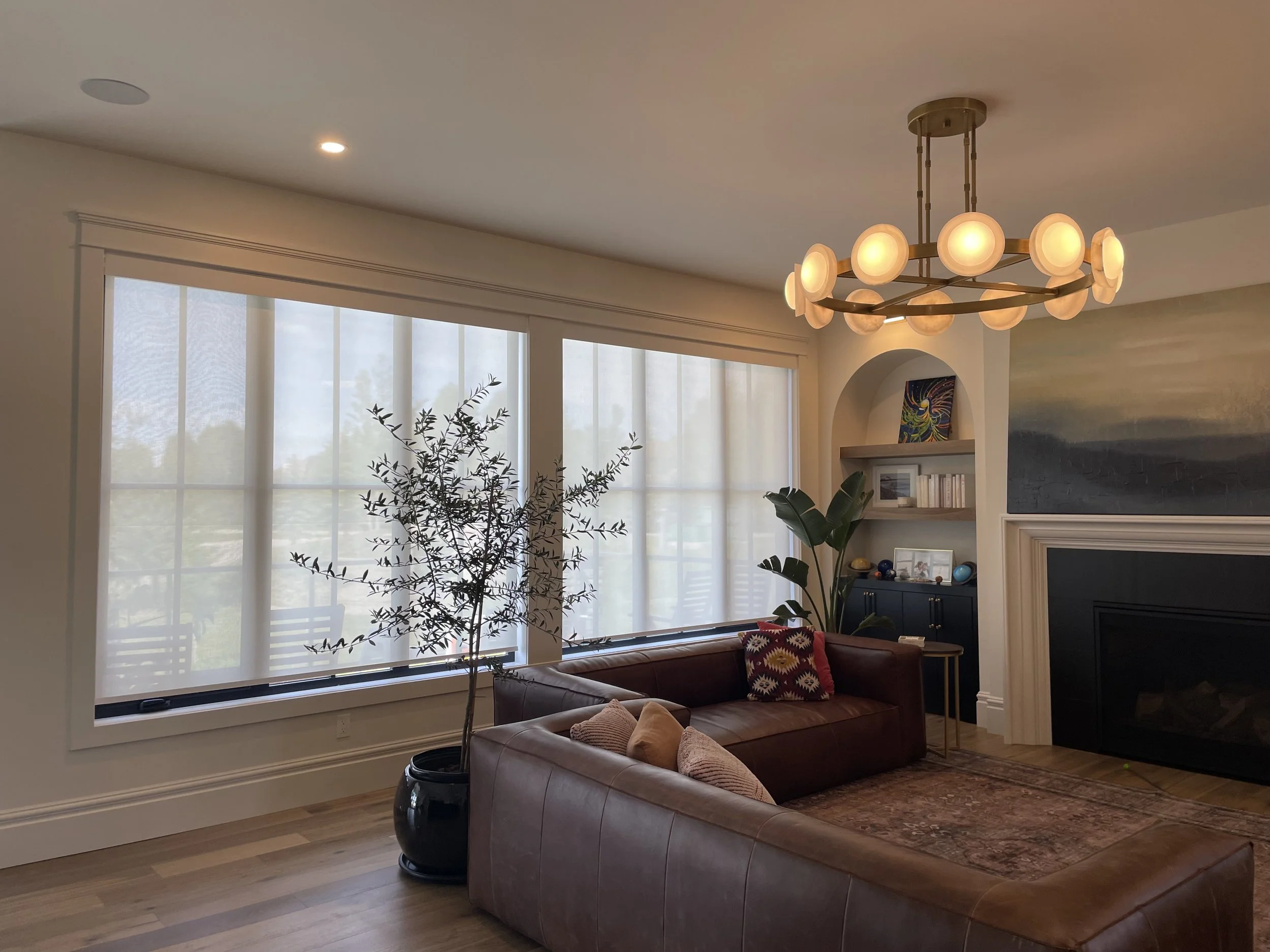 Living room with large windows, leather sofa with pillows, potted plant, built-in shelves, abstract painting, and a modern chandelier.