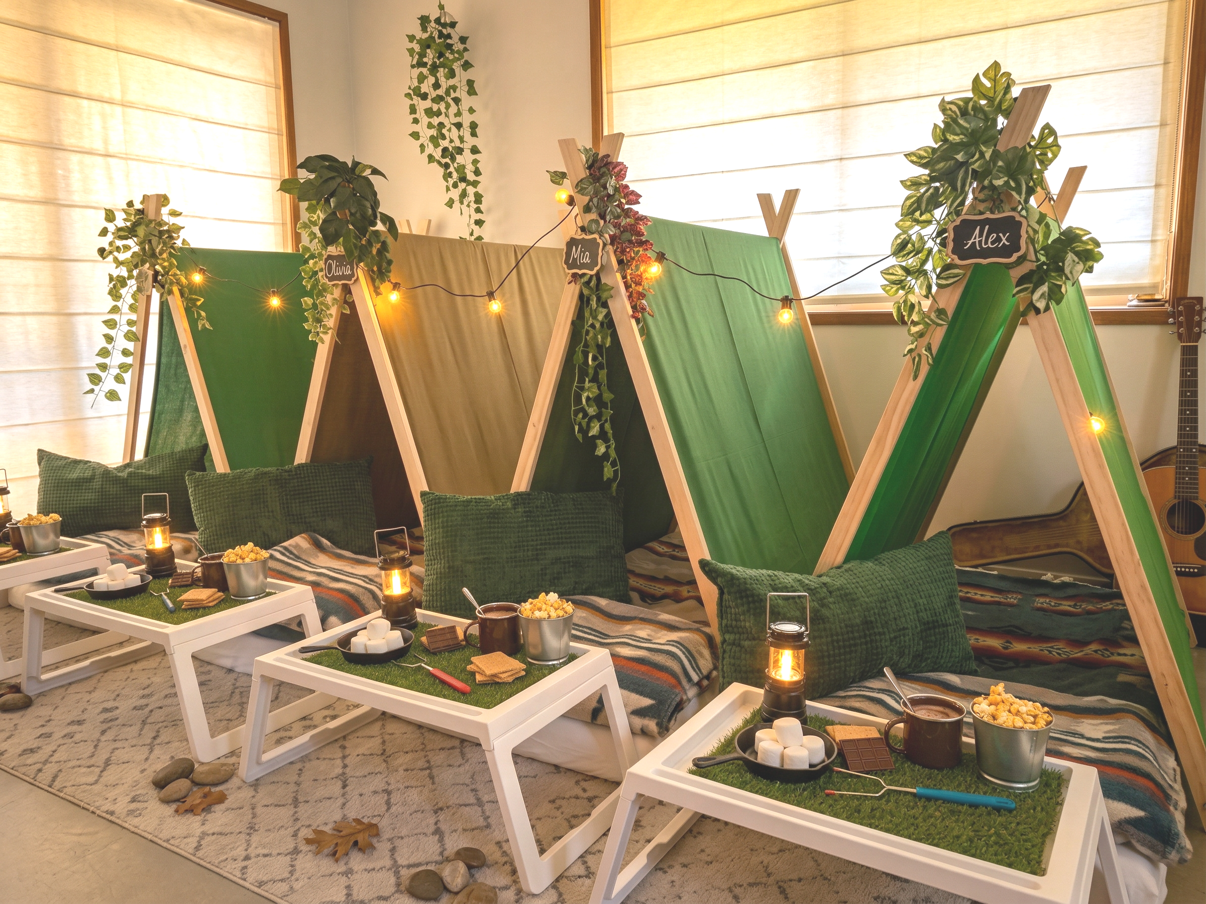 Cozy indoor camping setup with miniature tents, green pillows, string lights, and small tables with snacks and drinks.