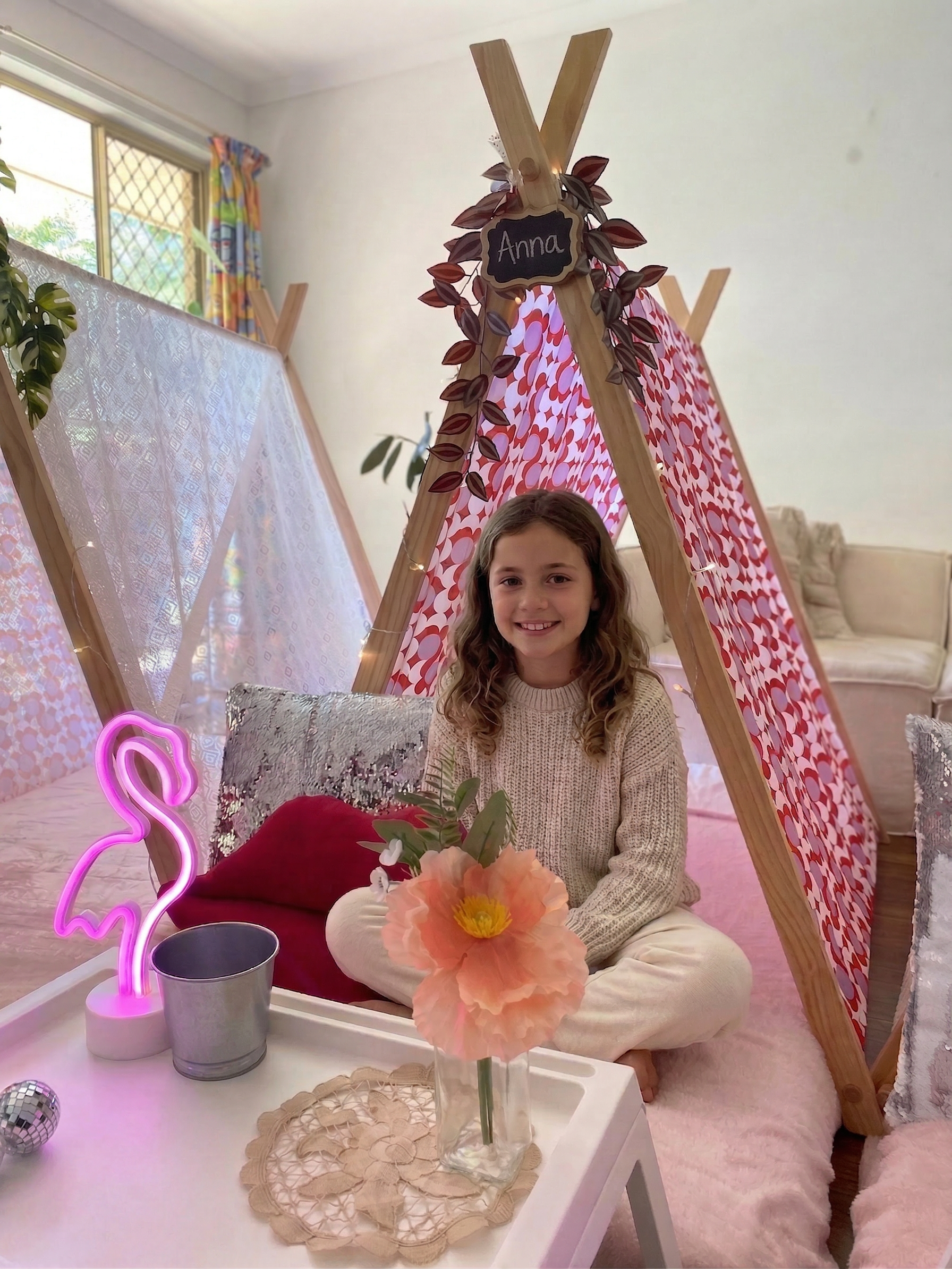 A young girl sitting cross-legged inside a pink and red patterned tent, smiling at the camera. The tent is decorated with leaves and a chalkboard sign that reads 'Anna.' In front of her is a small table with a vase of pink flowers, a neon pink flamingo light, a small metallic bucket, and a sequin pillow.