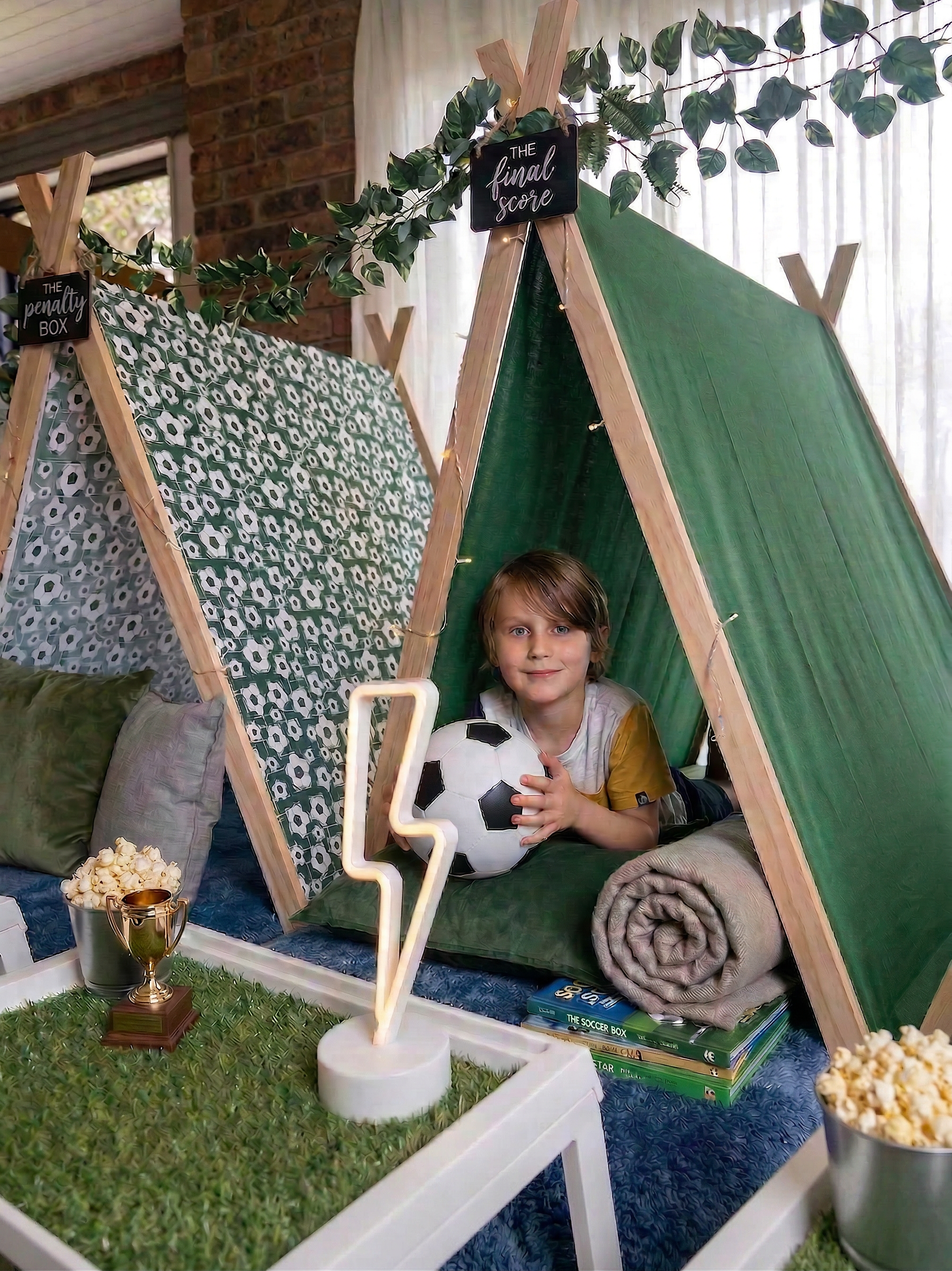Child inside a green and patterned teepee tent, holding a soccer ball, with books, rolled blanket, and decorative lights. Decor includes signs labelled 'The final score' and 'The penalty box,' along with greenery and fairy lights.