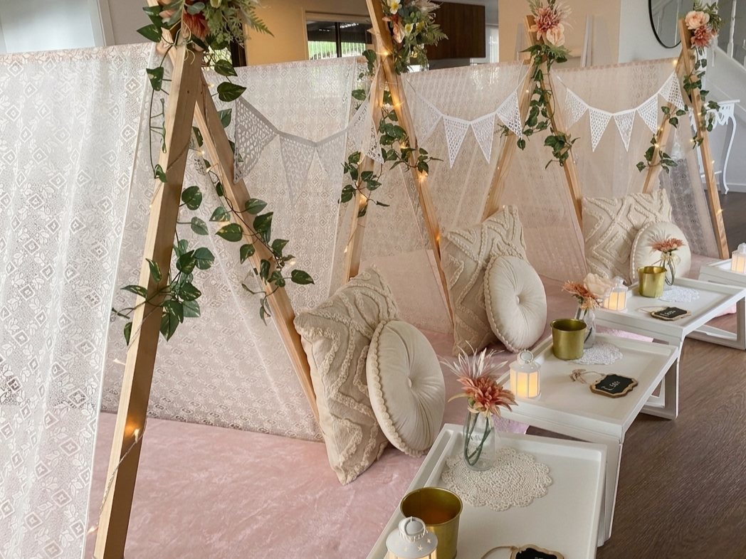 Cozy indoor seating area with wooden teepee tents decorated with lace, bunting, and fairy lights, featuring cushions, vases with flowers, candles, and small tables.