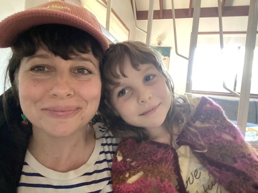 A woman and a young girl taking a selfie together indoors. The woman is wearing a pink cap and a striped shirt, while the girl has long curly hair and is wearing a colorful cardigan.