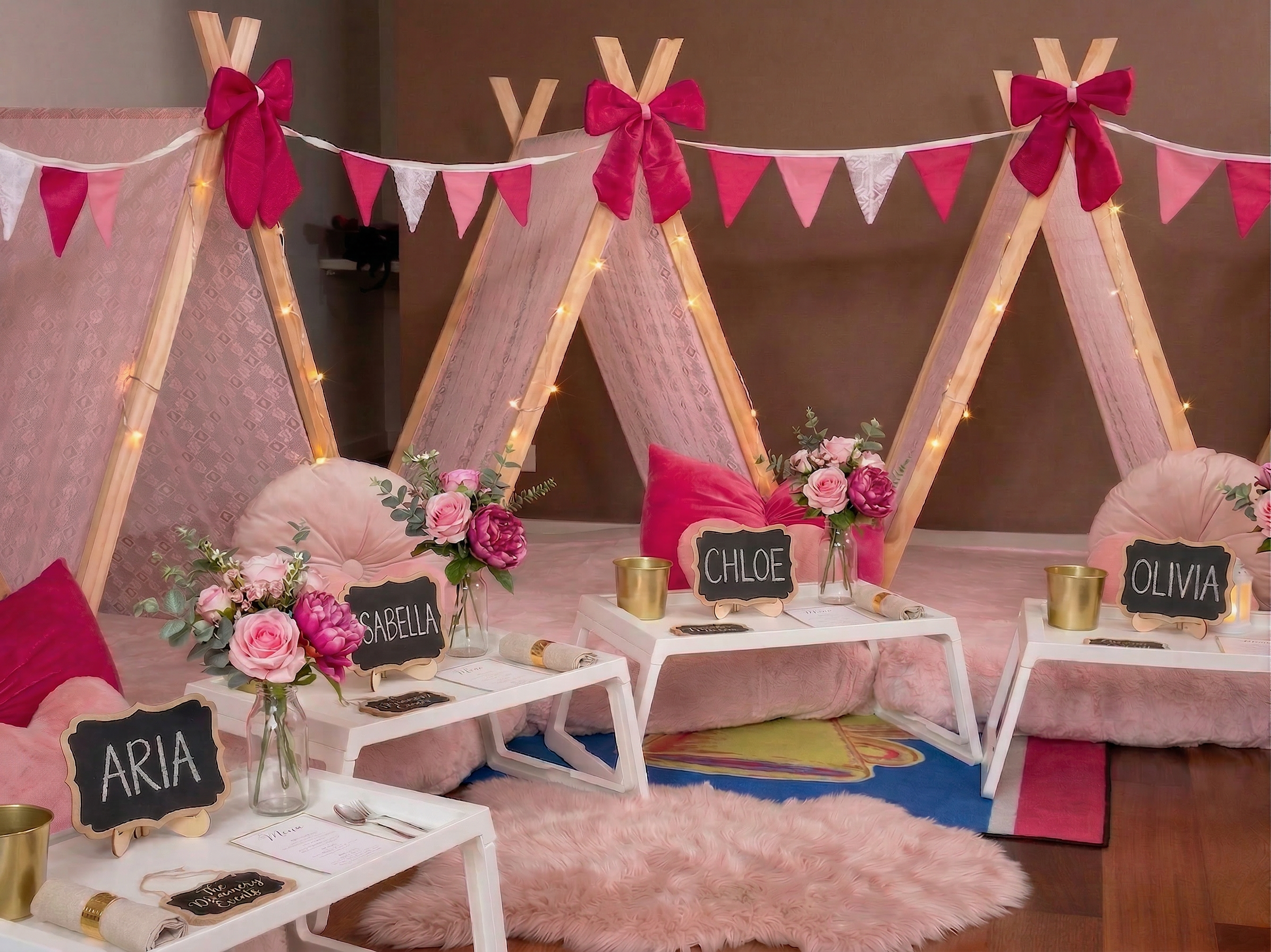 Decorative party setup with three small tables labeled with names 'Aria', 'Isabella', 'Chloe', and 'Olivia', topped with pink and purple flower arrangements, pink and white cushions, lamps, chalkboard signs. Behind is a teepee with pink, white, and red bunting, and string lights, on a pink fluffy rug.