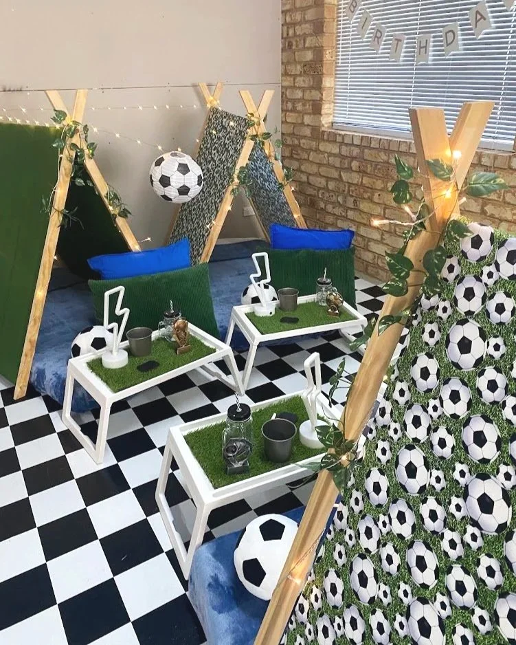 Child inside a green and patterned teepee tent, holding a soccer ball, with books, rolled blanket, and decorative lights. Decor includes signs labelled 'The final score' and 'The penalty box,' along with greenery and fairy lights.