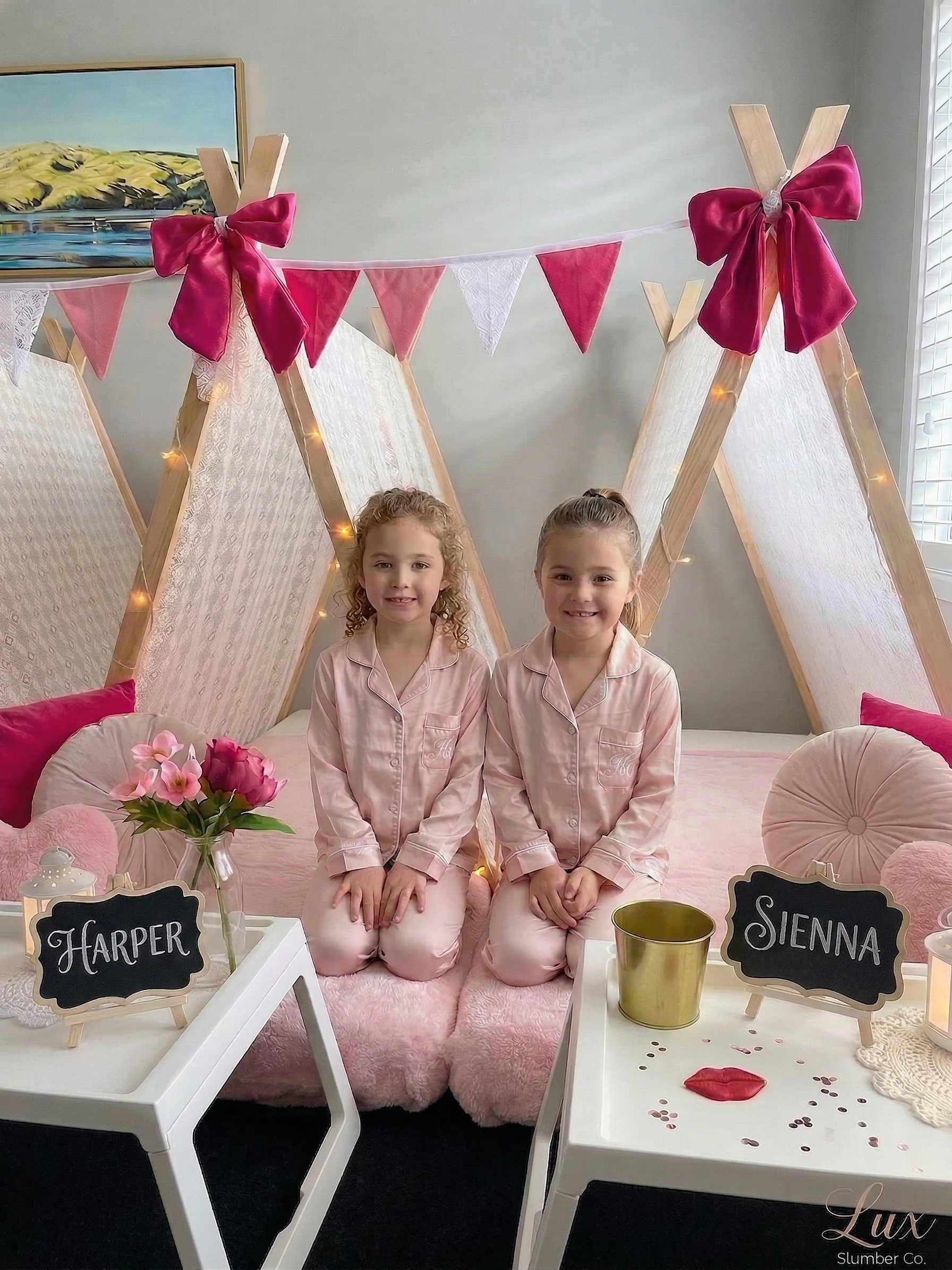 Two young girls in matching pajama sets sitting on a pink fluffy bed with teepee tents decorated with pink bows and bunting, surrounded by flowers and personalized chalkboard signs, celebrating a slumber party.