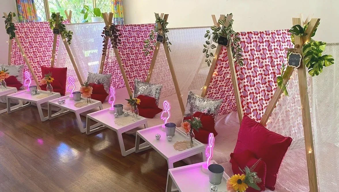 A cozy indoor space with teepee-style fabric tents decorated with pink and red patterned fabrics, supported by wooden frames, with greenery and string lights adorning the top, and small white tables in front of each tent holding vases, candles, and neon flamingo lights, decorated for a celebration or gathering.