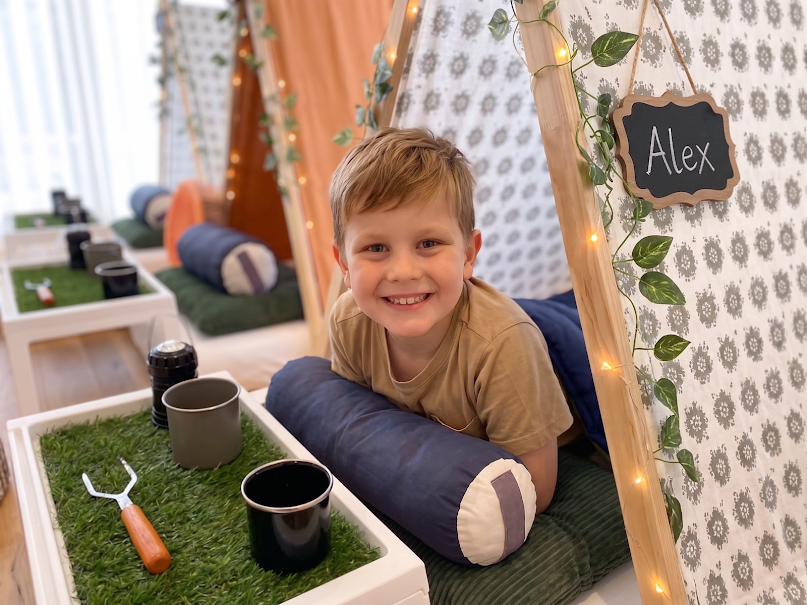 Indoor cozy camping scene with three teepees, each labeled with a name on a small chalkboard. The teepees are decorated with green and beige fabric,fake vines, and string lights. In front of each teepee are small white tables with snacks, marshmallows, and hot chocolate. There are green pillows inside. The floor has a carpet with scattered leaves and stones. A guitar is seen in the background near the window.