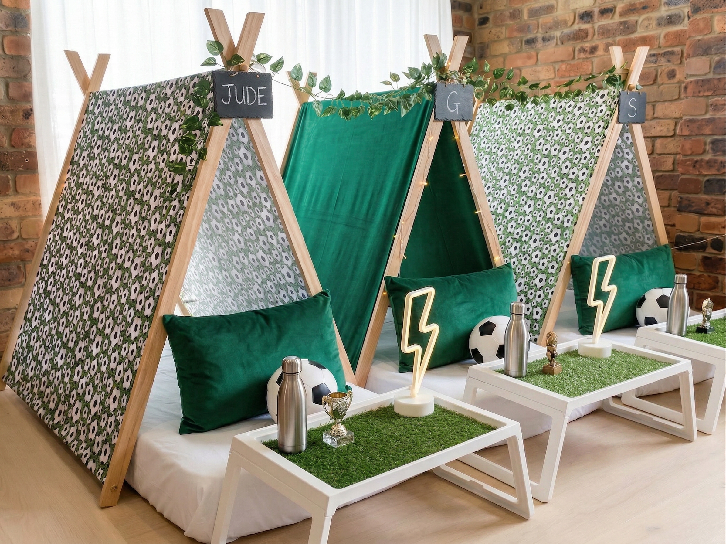 Indoor setup of three small tents with wooden frames, decorated with green pillows, footballs, and metallic water bottles, labeled with chalk signs for Jude, G, and S, with small tables in front with trophies and lightning bolt light decorations.