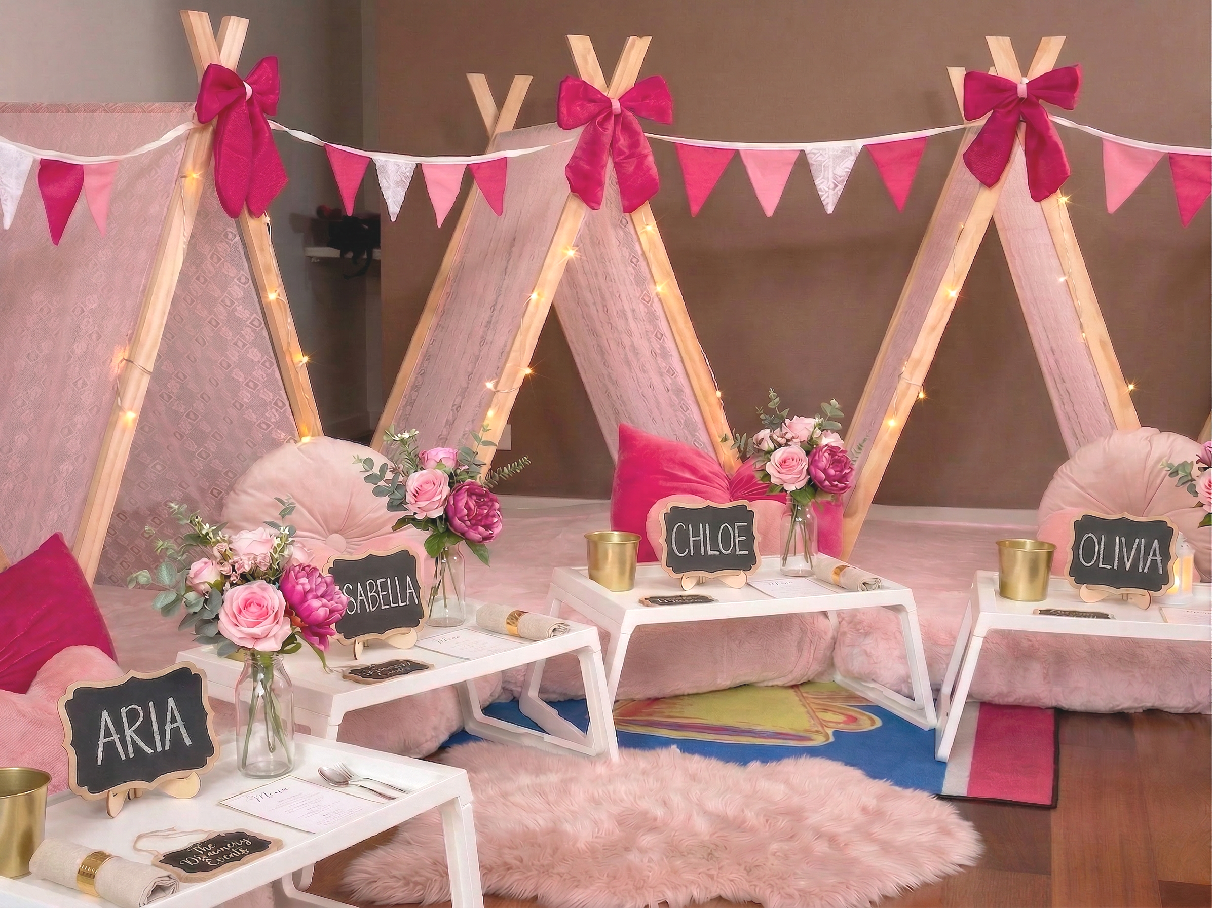 Decorative party setup with three small tables labeled with names 'Aria', 'Isabella', 'Chloe', and 'Olivia', topped with pink and purple flower arrangements, pink and white cushions, lamps, chalkboard signs. Behind is a teepee with pink, white, and red bunting, and string lights, on a pink fluffy rug.