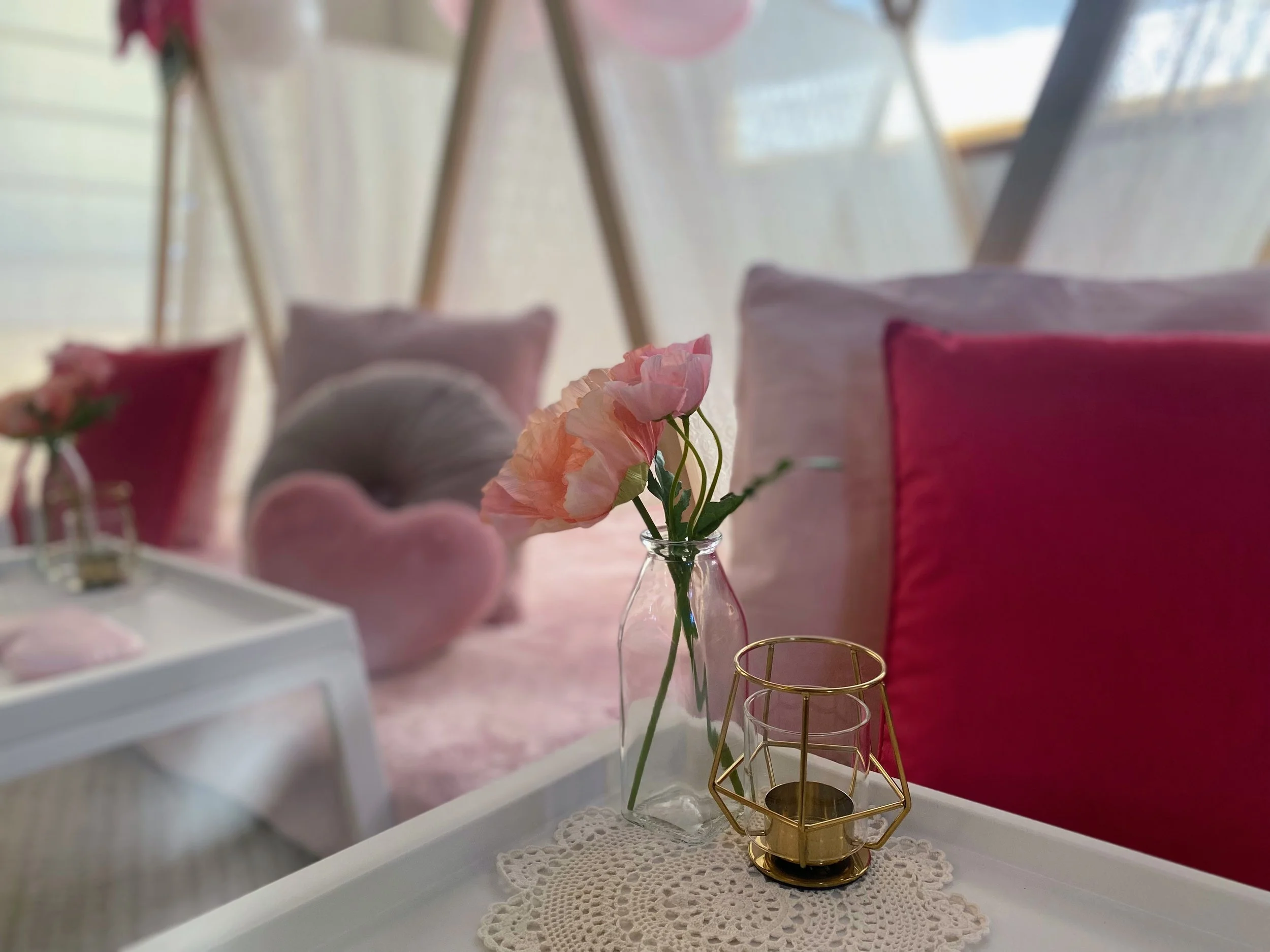 Close-up of a small white table with a Moroccan-style candle holder, a glass vase with pink flowers, and a lace doily. In the background, there are pink, gray, and white pillows and plush cushions on a soft pink surface. Blurred background of a cozy, pastel-colored room.