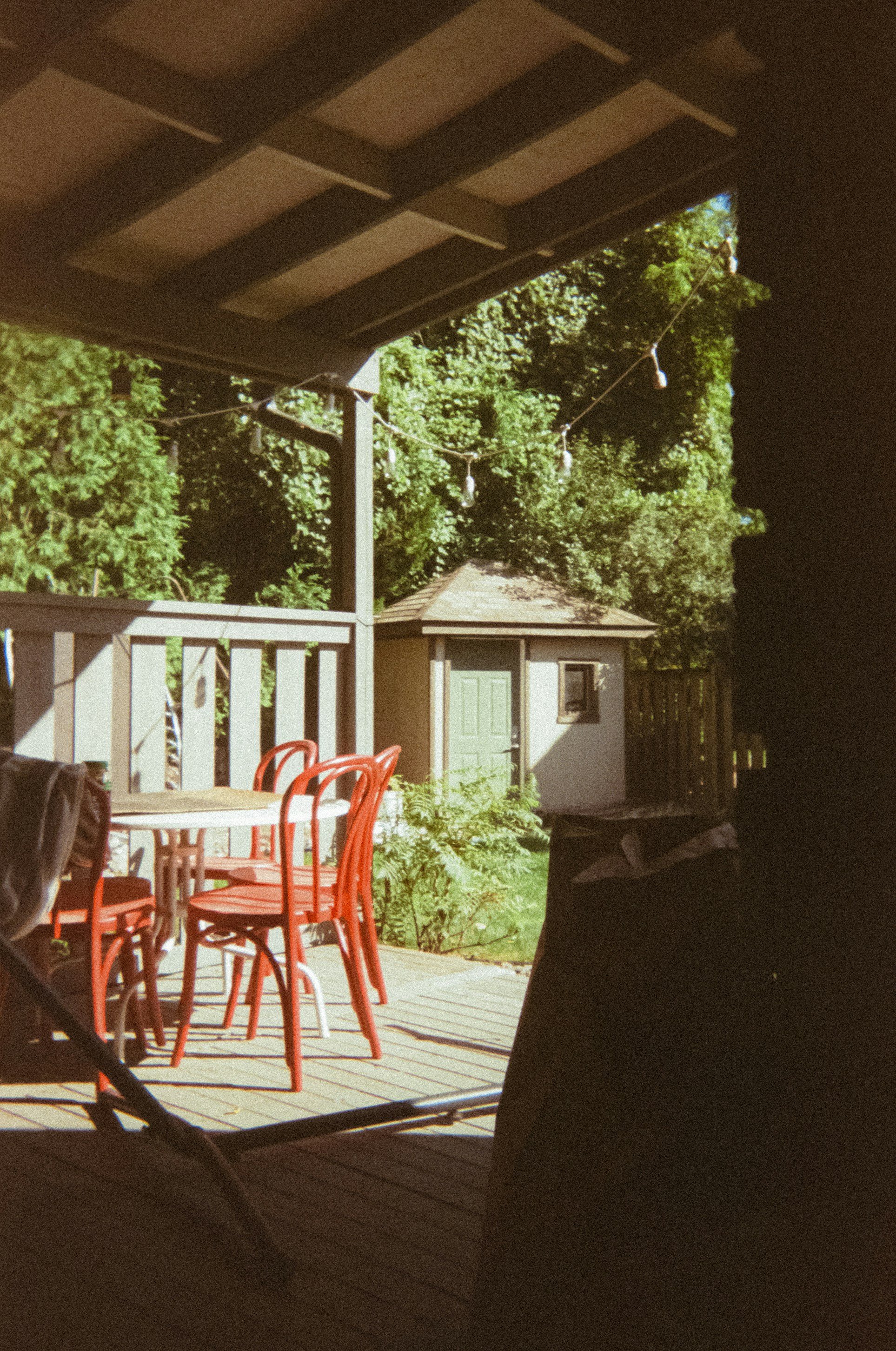 Backyard patio with red chairs and table, string lights, greenery in background, and a small shed.