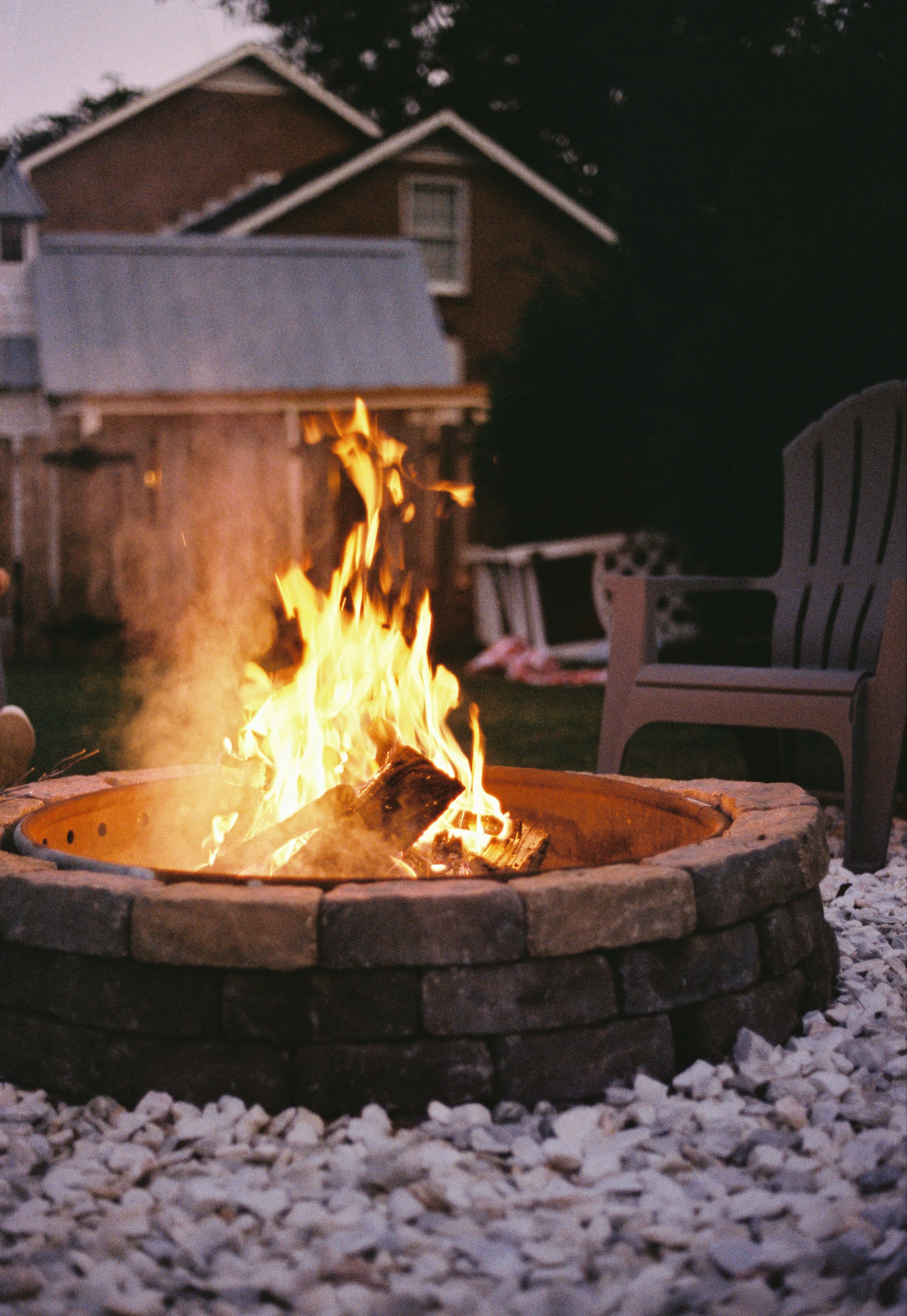Outdoor fire pit with burning logs, surrounded by stone, in a backyard setting with a house and chair visible.
