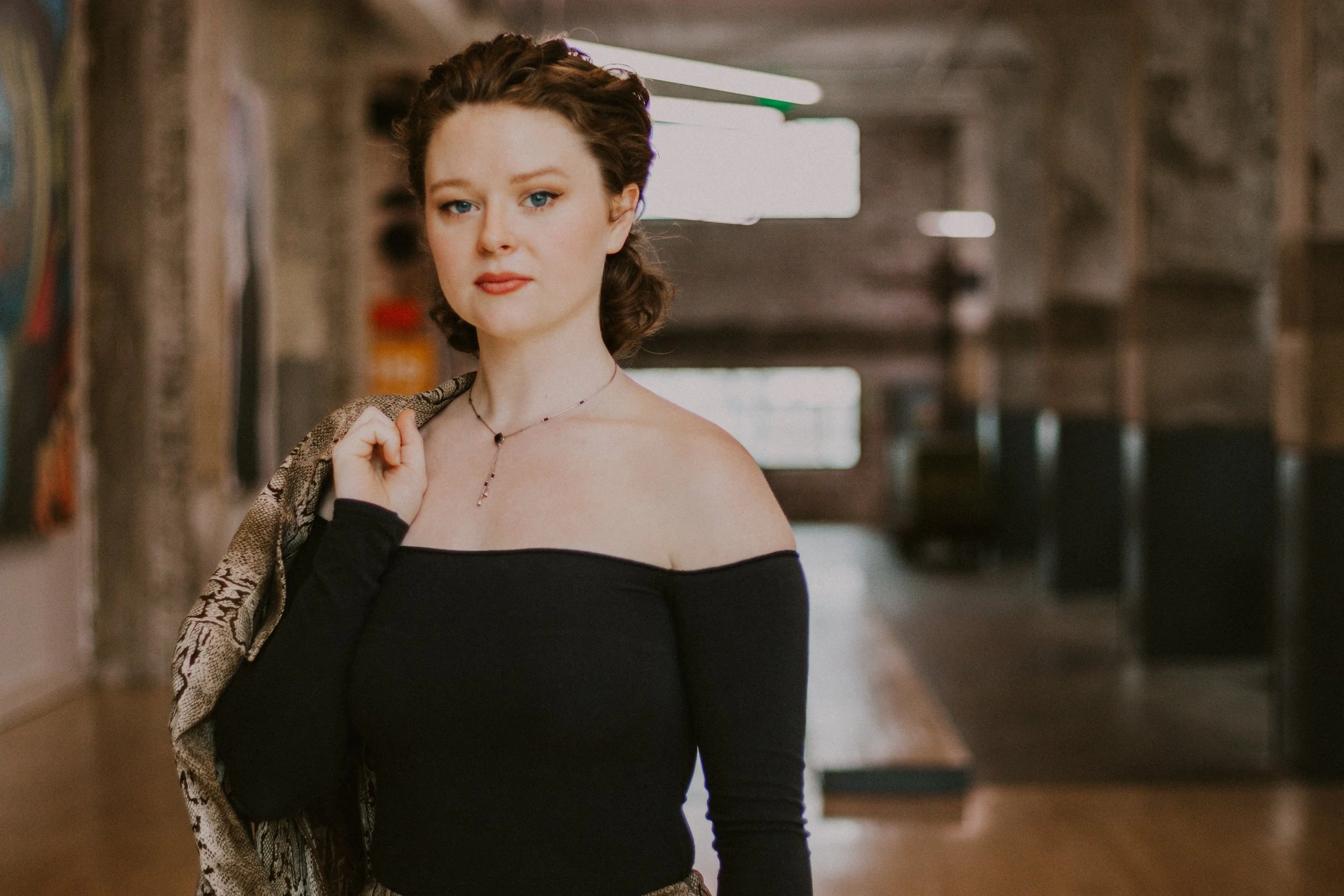 Sarah Neal with wavy brown hair and fair skin standing in Crosstown Concourse with artwork on the walls, wearing an off-the-shoulder black top, holding a jacket over her shoulder, and wearing a delicate necklace.