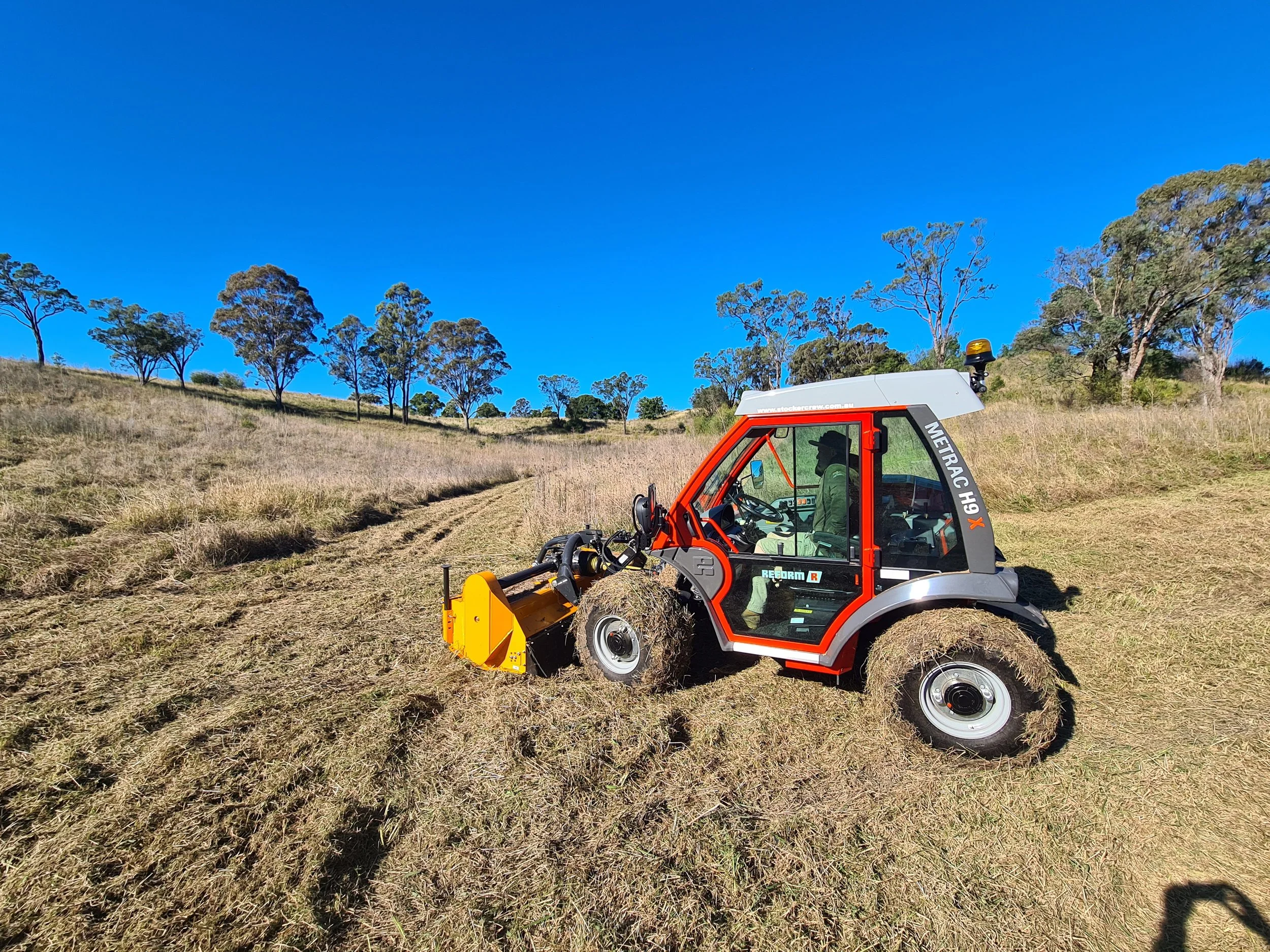 A red and silver utility vehicle, labeled "METRAC H9X," with a driver, operating on a mowed grassy field with trees in the background under a clear blue sky.